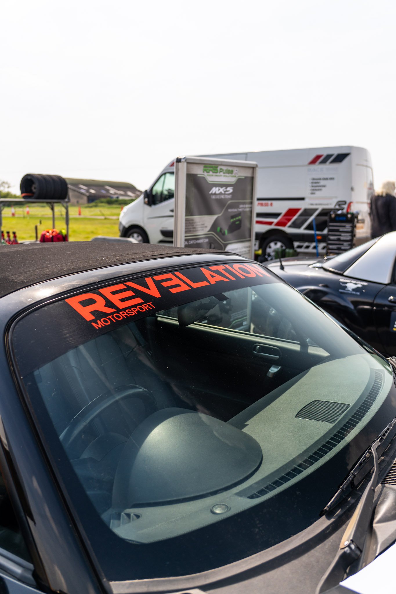 Close-up of a car windshield with a red 'REVELATION' decal, parked in a lot with other vehicles and racing setup equipment visible in the background.