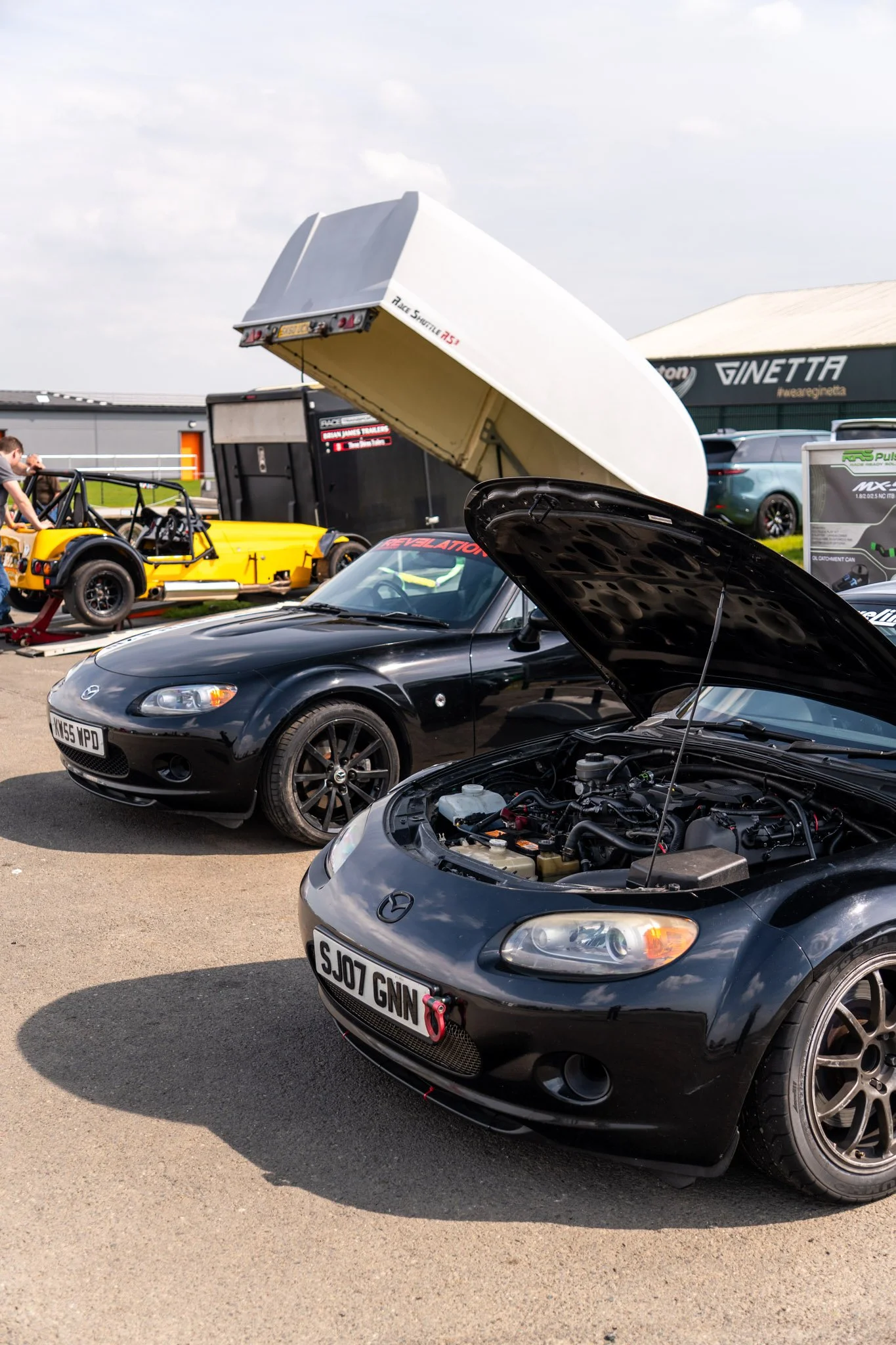 Multiple cars, including a black Mazda MX-5 with its hood open and a yellow race car on a trailer, are present at a car event, with a building labeled Ginetta and some vehicles in the background.