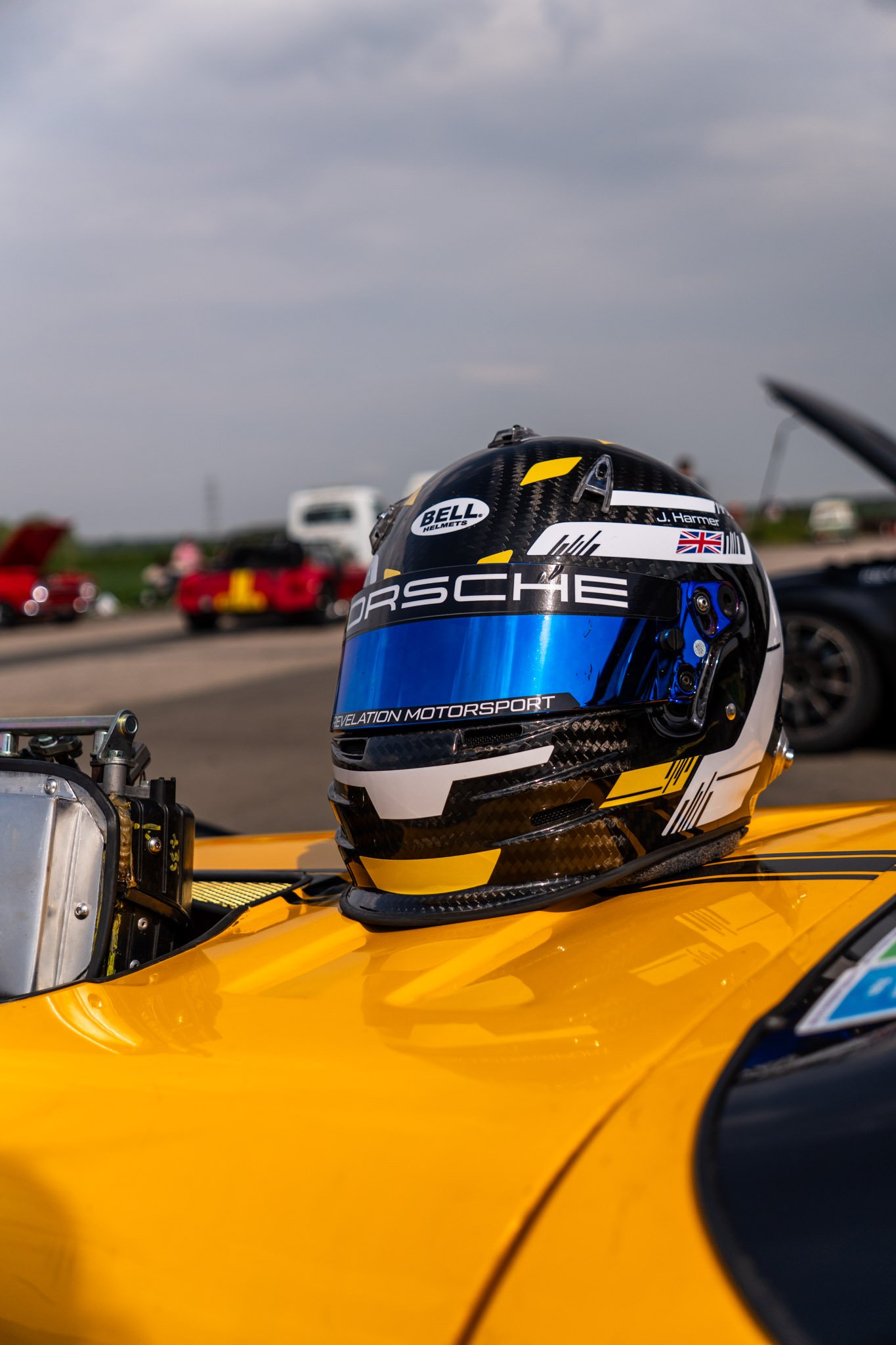 Close-up of racing helmet placed on a yellow race car, with a blurred background showing cars and a cloudy sky.
