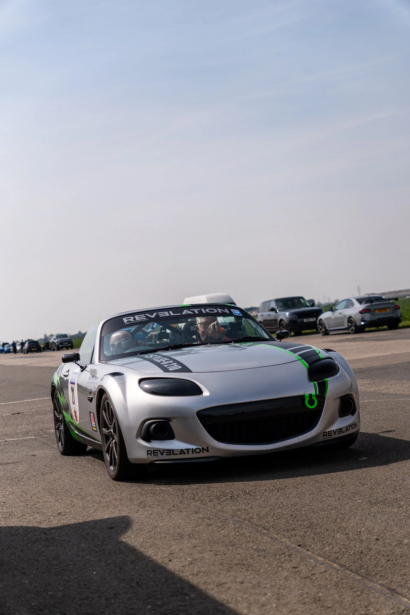 A silver car with a black and green racing stripe and the word 'Revelation' on the front bumper is parked on an open paved area under a partly cloudy sky. Multiple other cars are visible in the background, along with some people.