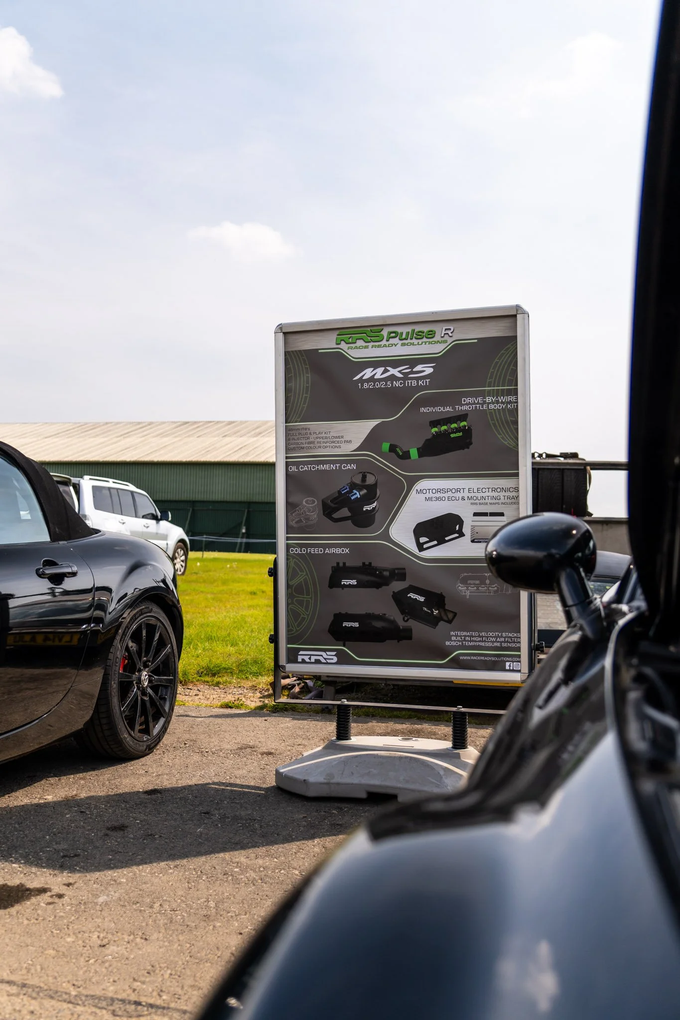 A large outdoor sign displays automotive racing parts and electronics, surrounded by parked cars and a grassy area with a building and a cloudy sky in the background.