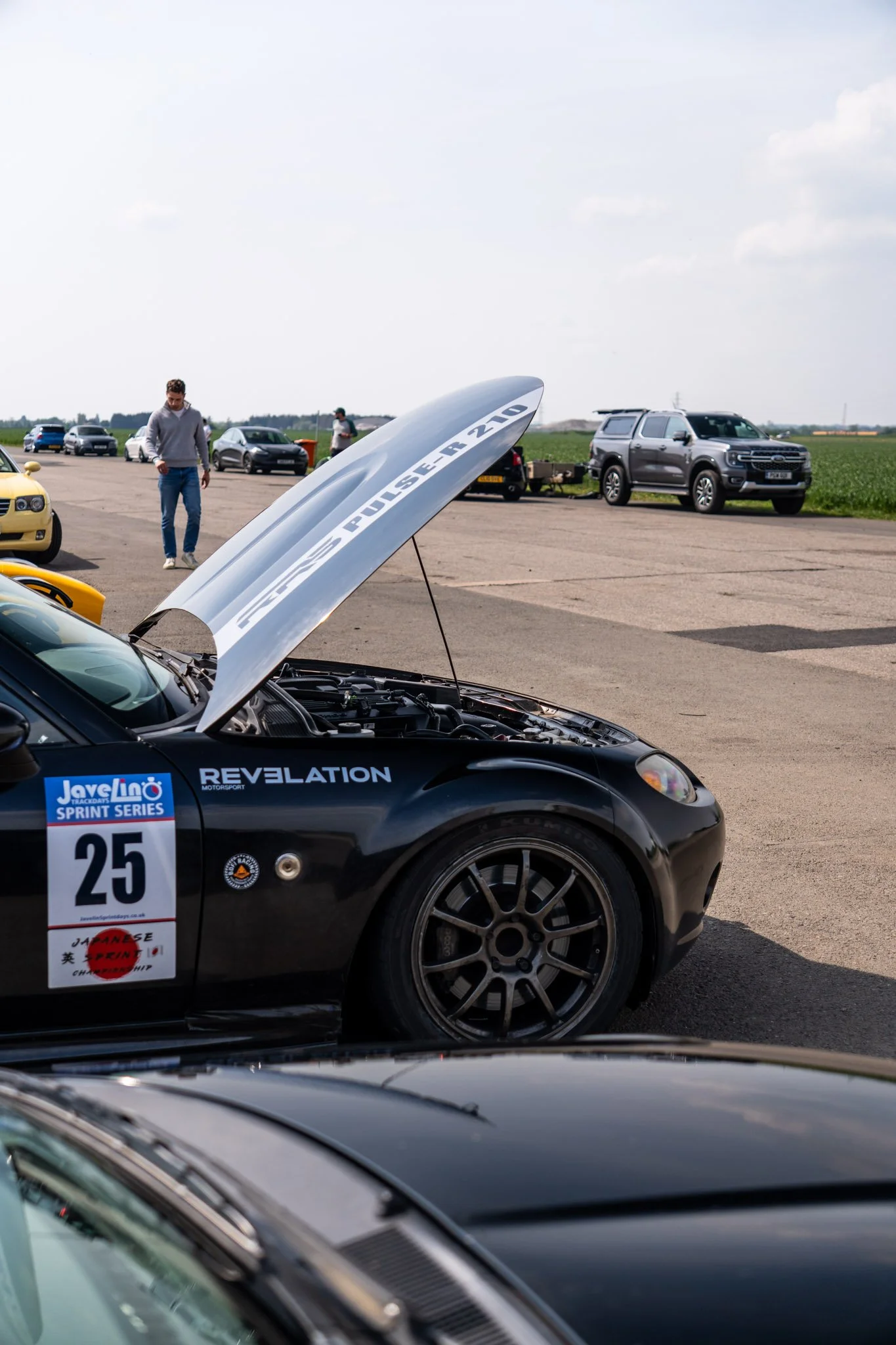 A black race car with sports decals and the number 25 on the side, parked outdoors at a racing event. The hood is open, revealing the engine. There are people walking around and several other cars parked in the background, along with open fields and 
