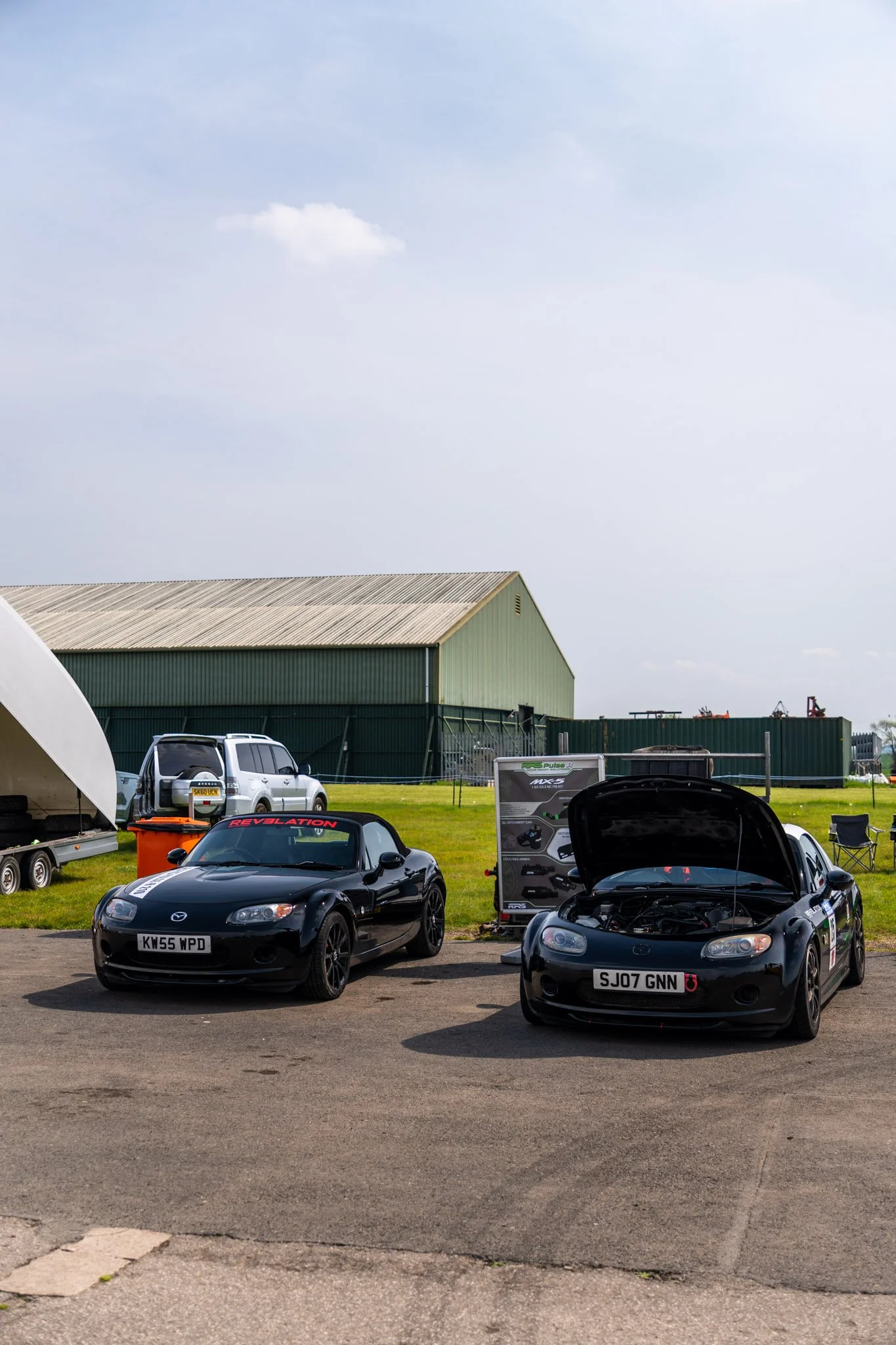 A car meet-up in a parking lot with two black Mazda MX-5 Miata sports cars in the foreground, one with its hood open. Several other cars, including a silver SUV, are parked nearby. There is a large green warehouse building in the background with a cl
