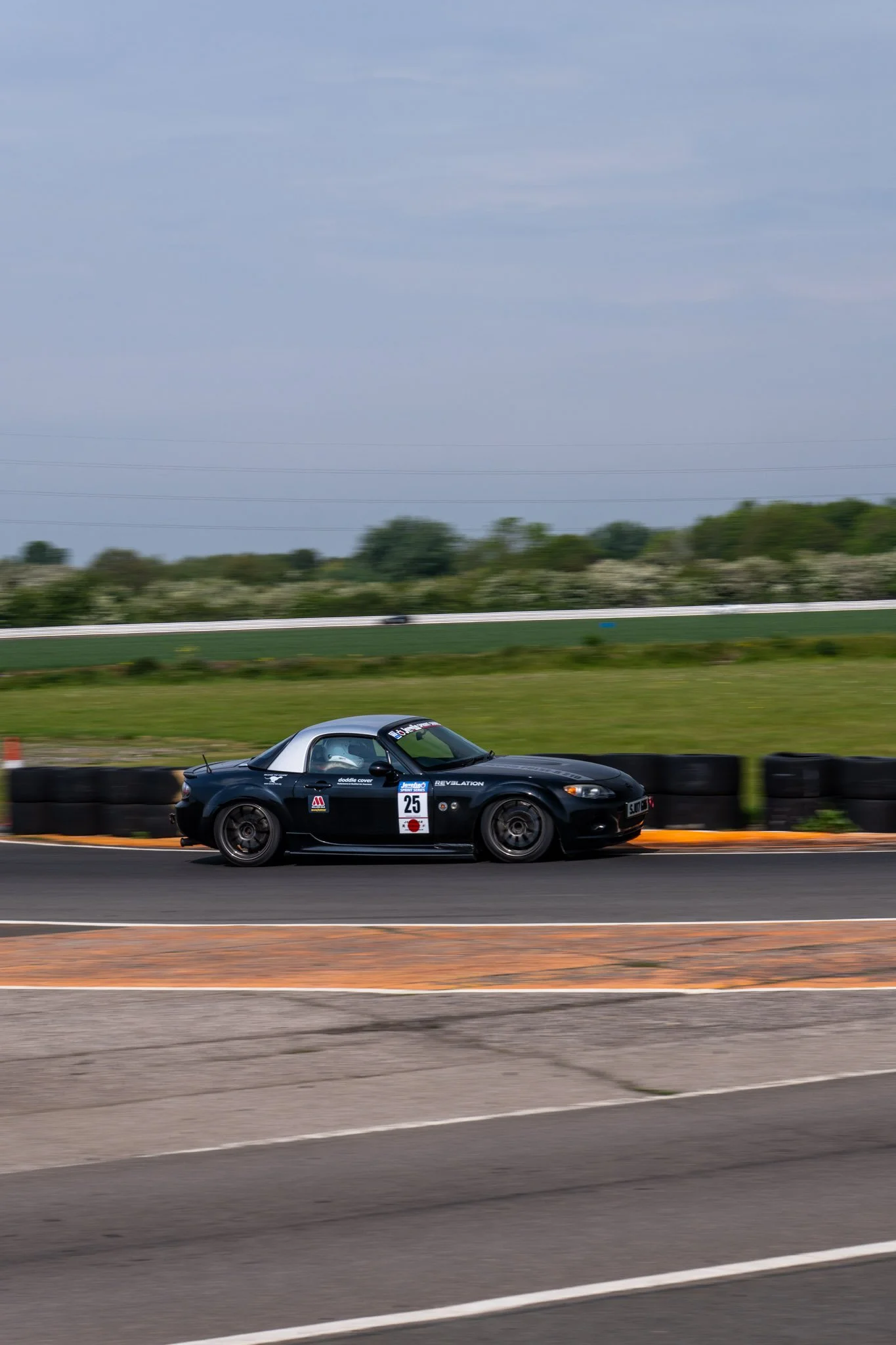 A black Mazda MX-5 Miata race car with the number 25 on the door, racing on a track with grass and trees in the background under a cloudy sky.