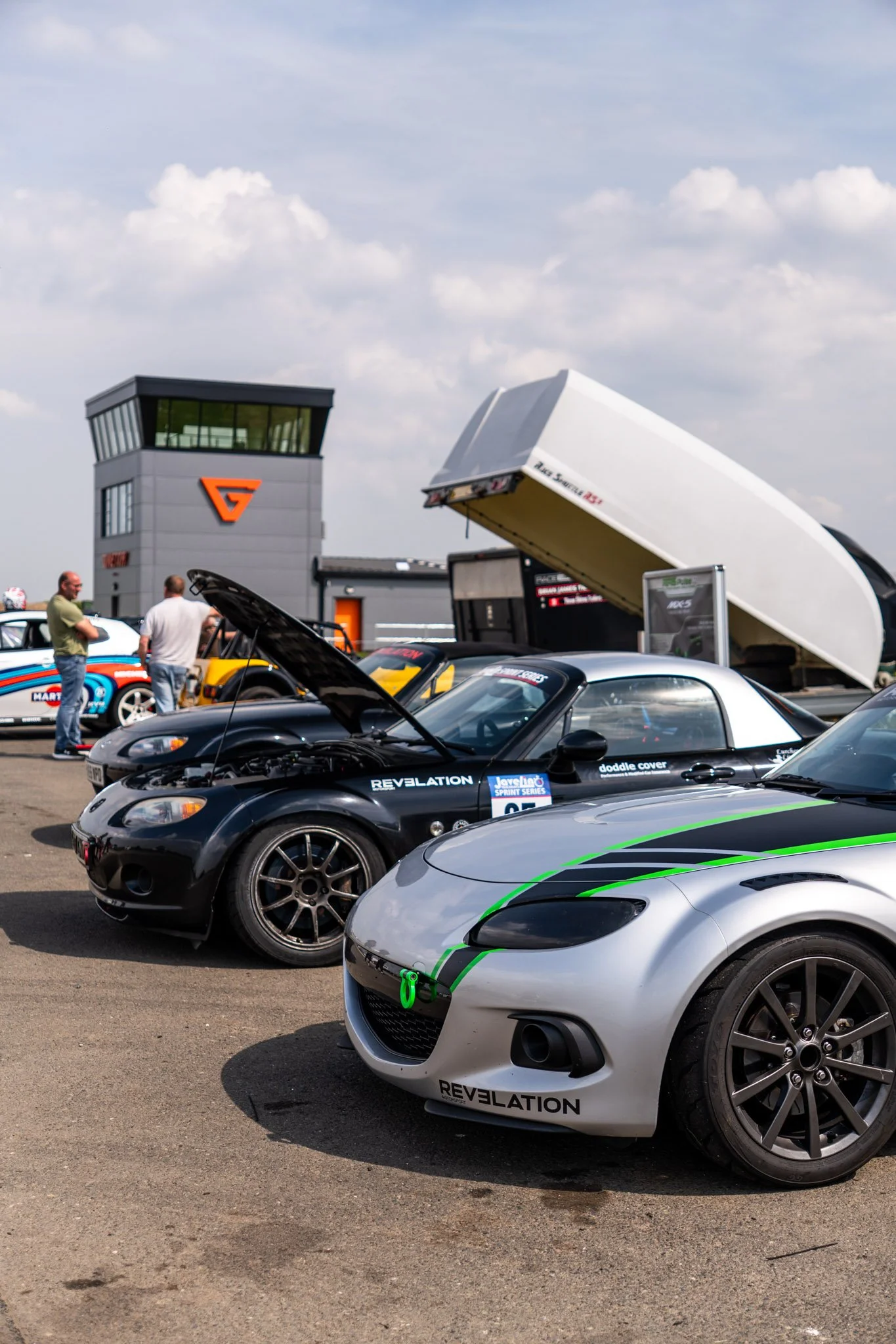 Multiple racing cars parked on a track with a control tower and a white aircraft in the background, during daytime with partly cloudy skies.