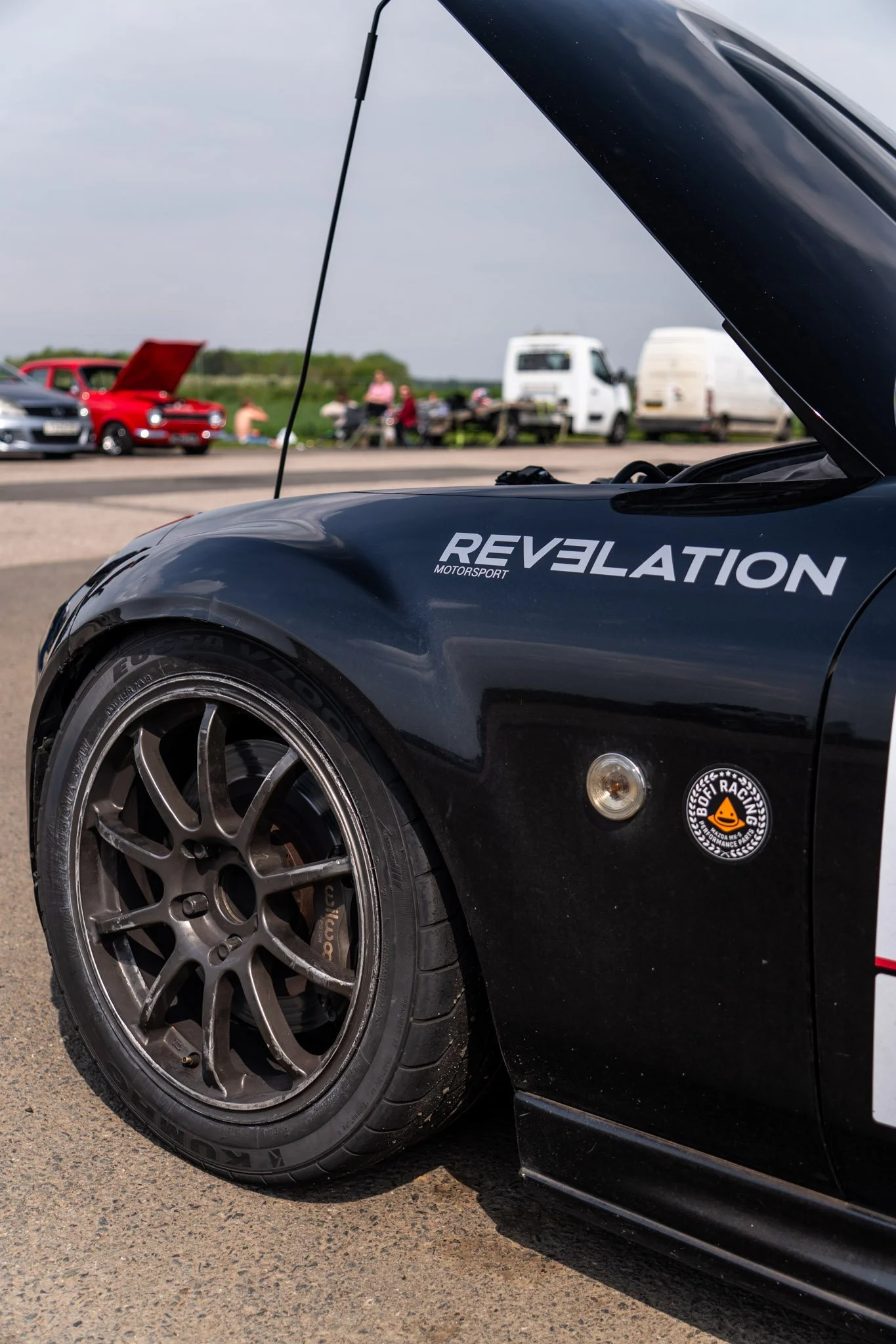 Close-up of a black sports car with a large rear tire and the word 'Revelation' on the side, parked on a roadway. In the background, there is a blurred scene of people, a red car with its hood open, and white vans, possibly at a car event or race.