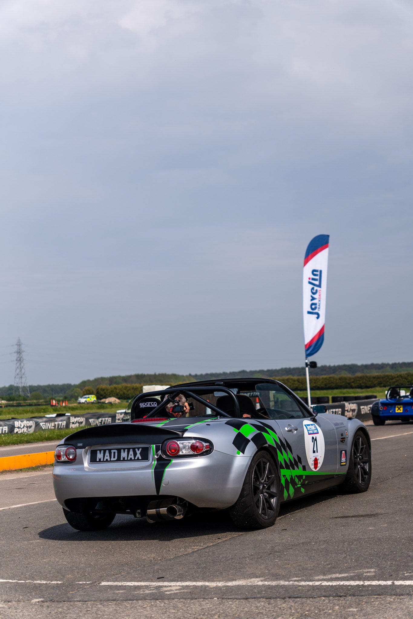 A silver sports car with a custom license plate reading 'MAD MAX' at a racing event, parked on pavement next to a yellow curb. The car has green and black racing graphics and a roll cage inside. In the background, there are other racing vehicles, a f