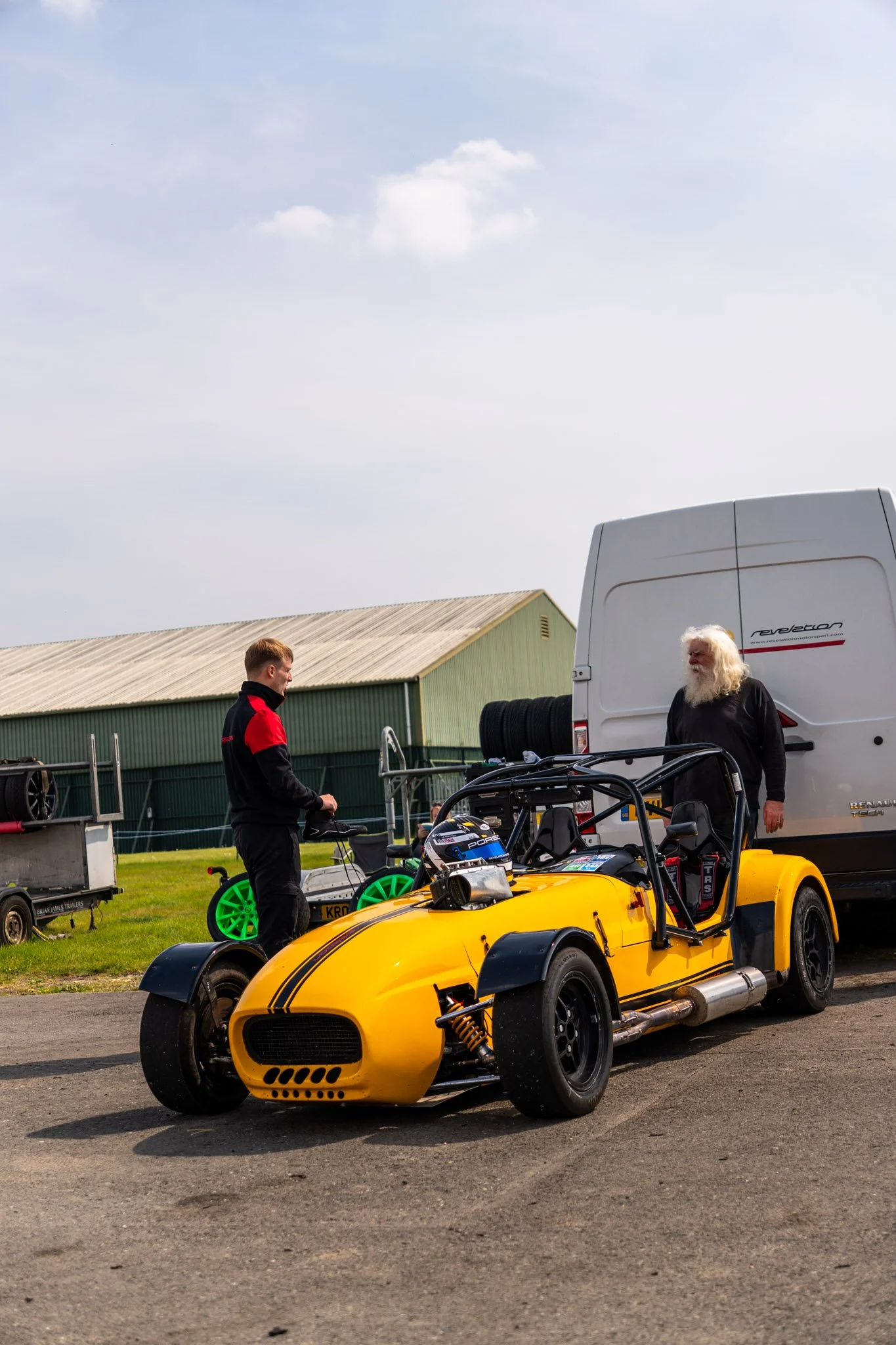 A yellow race car with a black stripe parked on a track, with two men near it, one in a black jacket with red accents and another with long white hair and beard. There are racing tires and other equipment around.