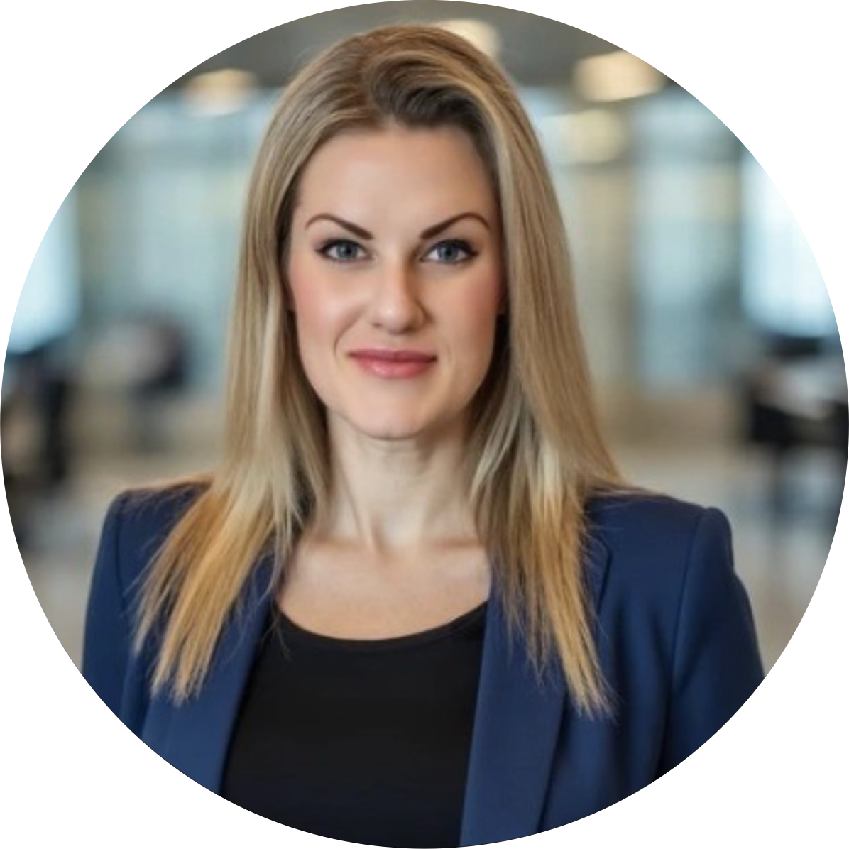 Professional woman with blonde hair smiling, wearing a navy blazer and black top, standing in a modern office environment.