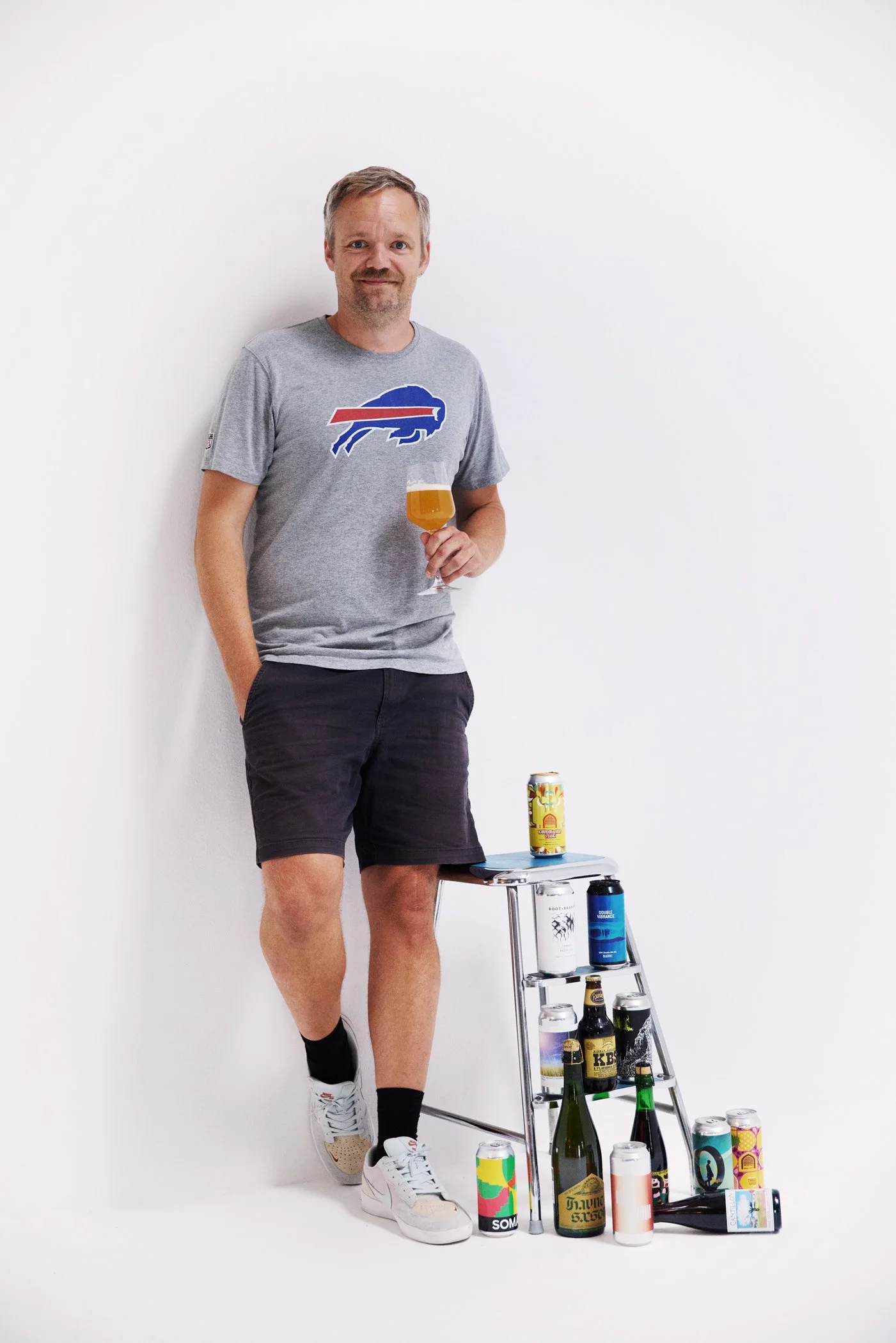 A man wearing a gray Buffalo Bills T-shirt, black shorts, and sneakers stands against a white background, holding a glass of beer, with a small metal shelf next to him filled with various beer bottles and cans.
