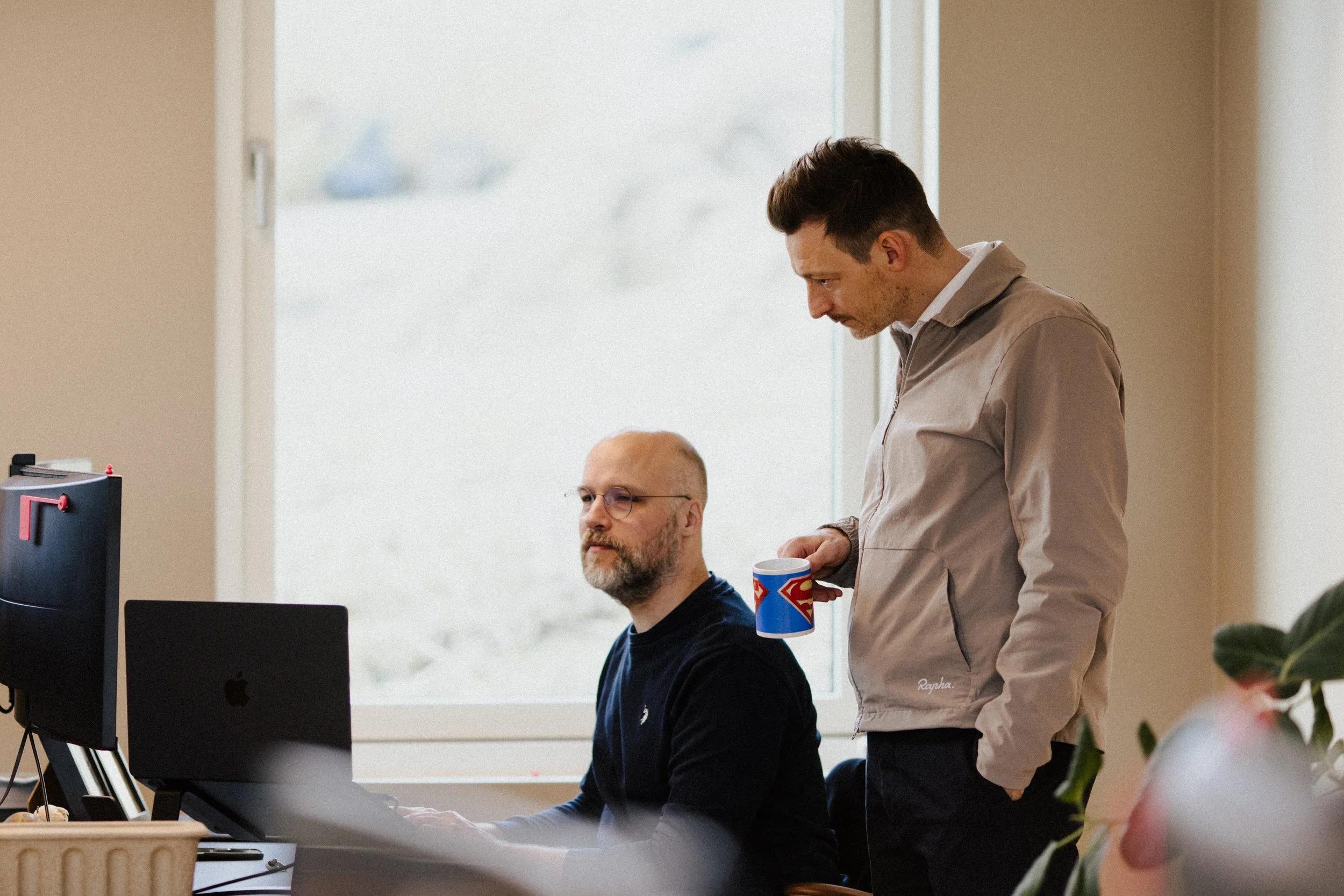 Two men in an office working on computers; one man is sitting at a desk with a MacBook and the other is standing next to him holding a Superman coffee mug.