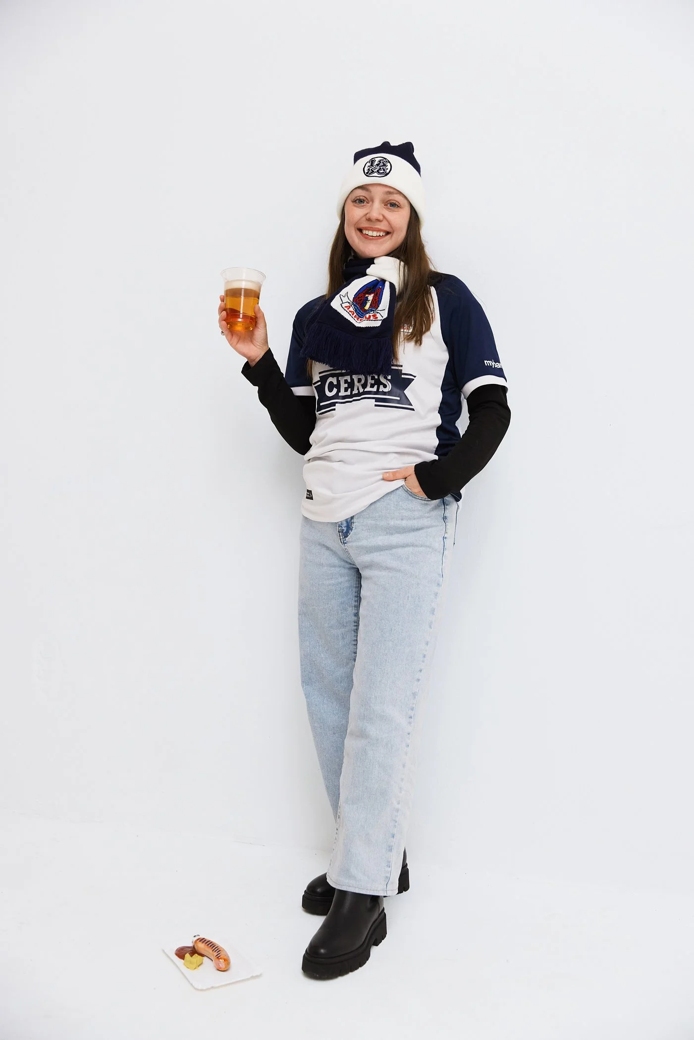 A woman wearing a sports jersey, scarf, and beanie hat stands against a plain white background, holding a drink in her right hand, smiling, with some sushi on a napkin on the floor in front of her.