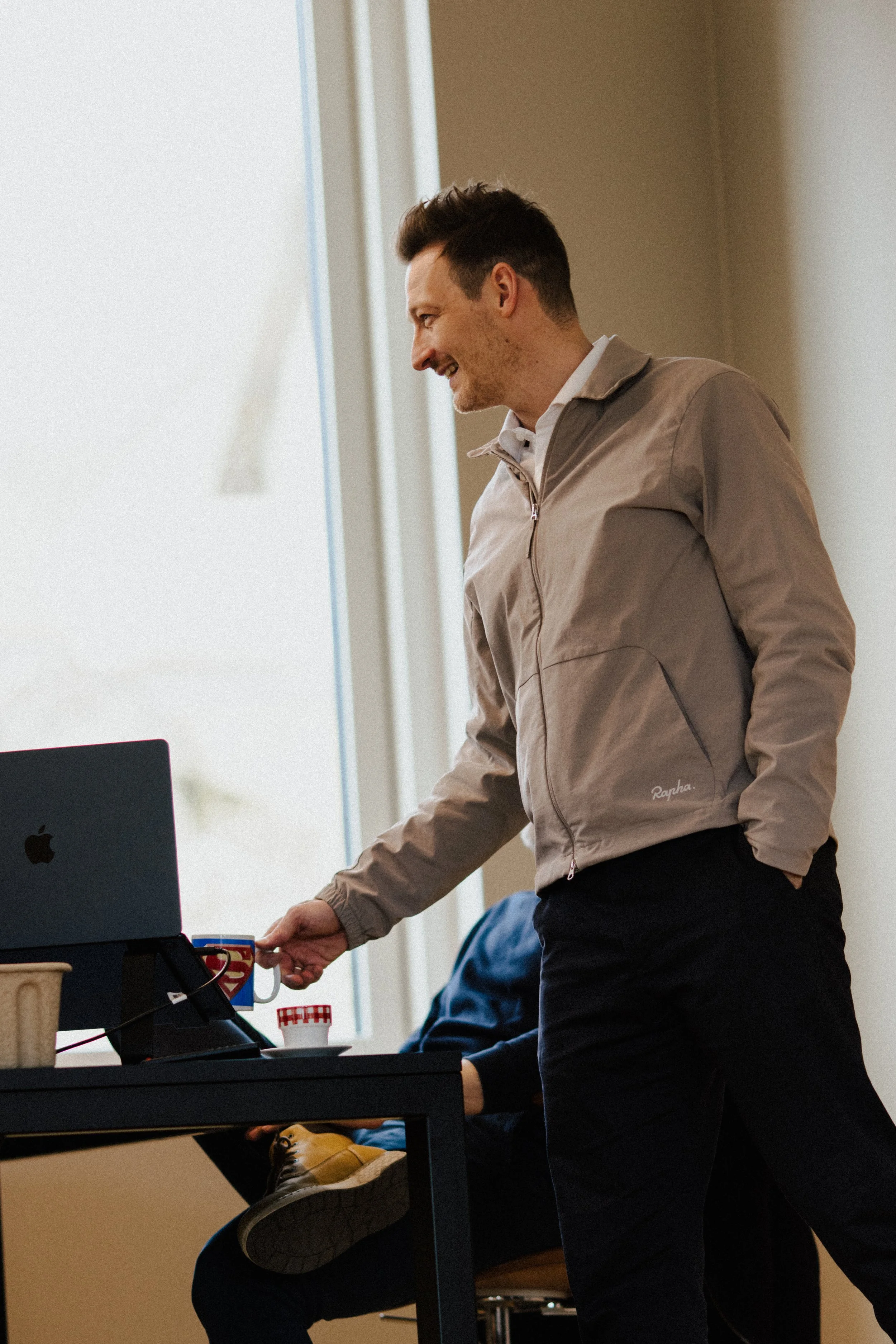 Man standing near desk, reaching for coffee mug, in a casual office setting.