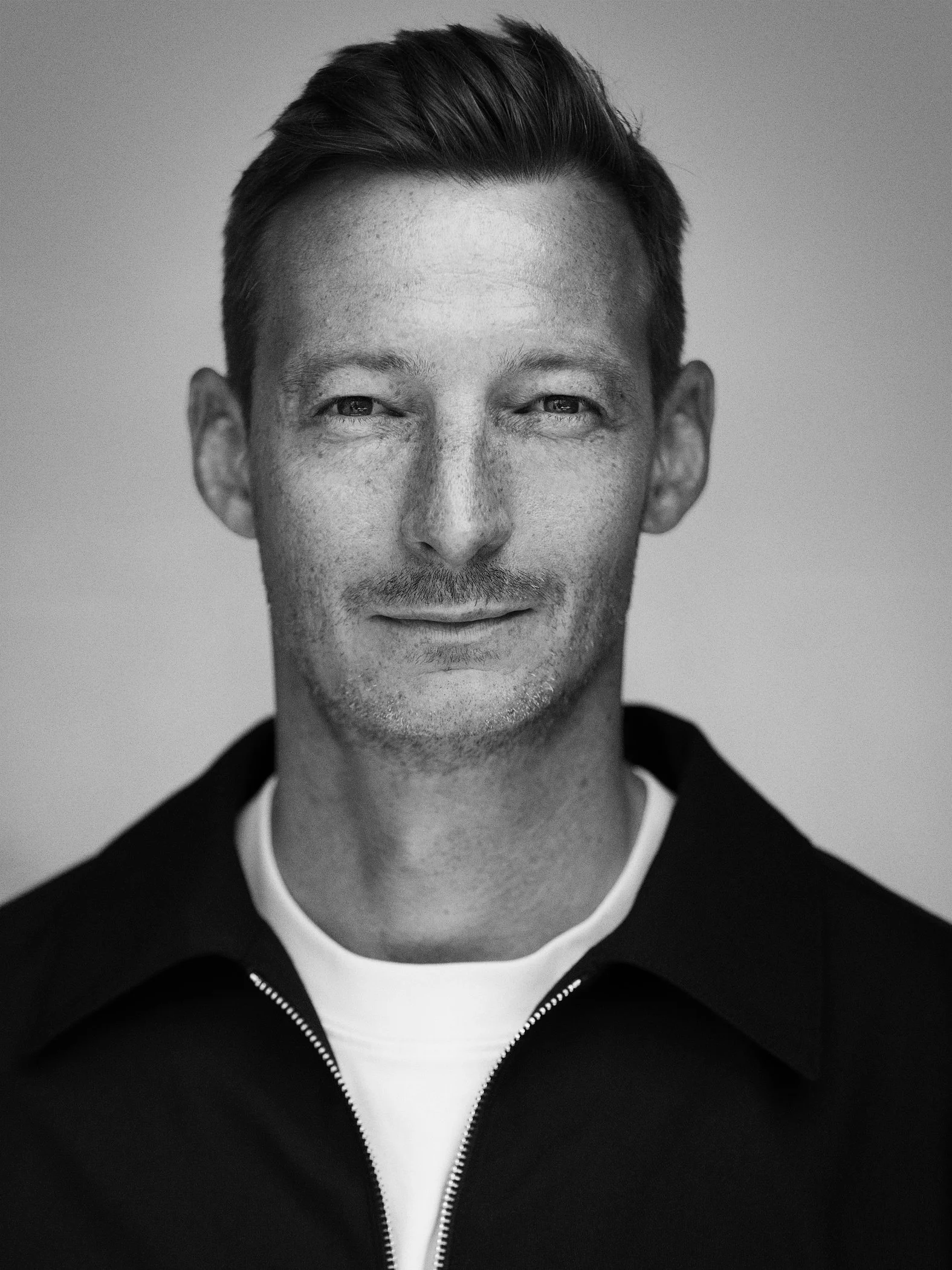 Black and white headshot of a man with short hair, slight beard, and a subtle smile. He's wearing a collared shirt with a zipper.