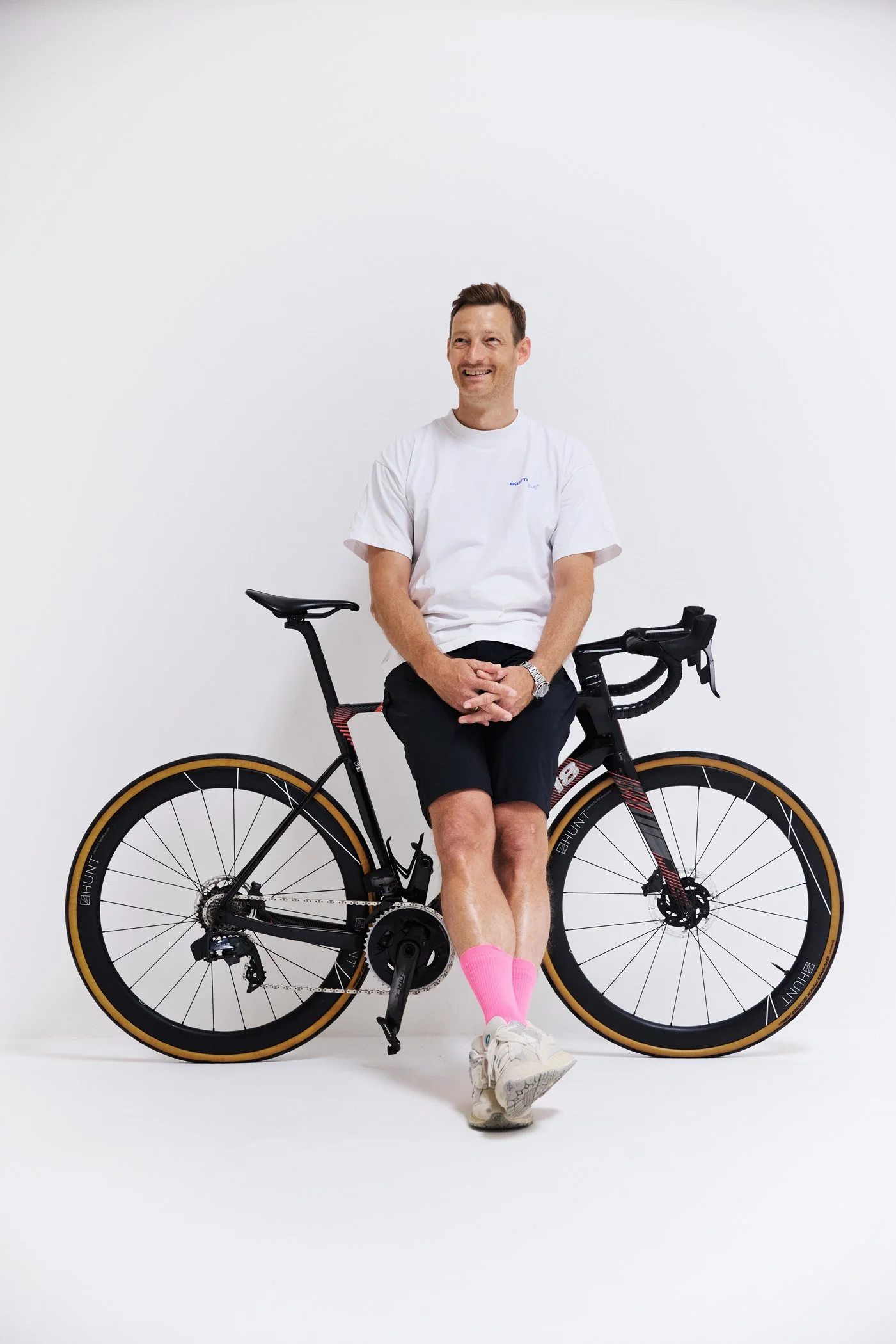 A man sitting on a bicycle against a plain white wall, wearing a white t-shirt, black shorts, pink socks, and sneakers, smiling.
