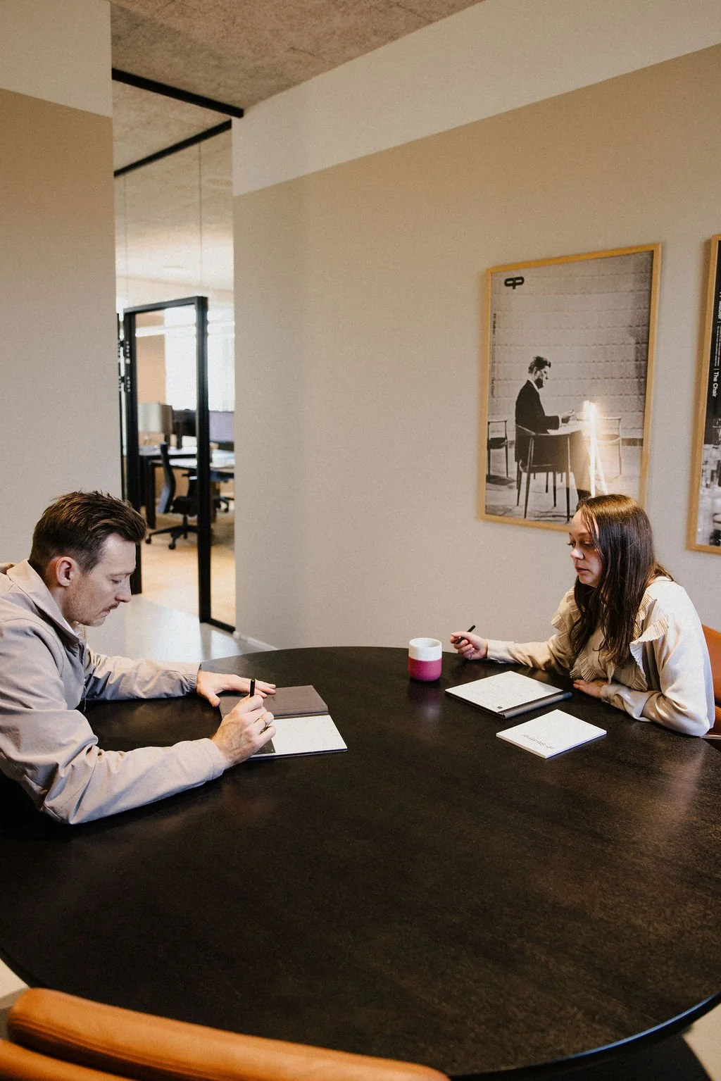 Two people sitting at a dark oval table in an office, taking notes and having a discussion. The man on the left has brown hair and wears a beige jacket, while the woman on the right has long dark hair and wears a cream-colored blouse. There are notebooks, pens, and a cup on the table, with framed black-and-white photographs of a woman working at a desk on the wall behind them. In the background, there are office rooms with glass doors.