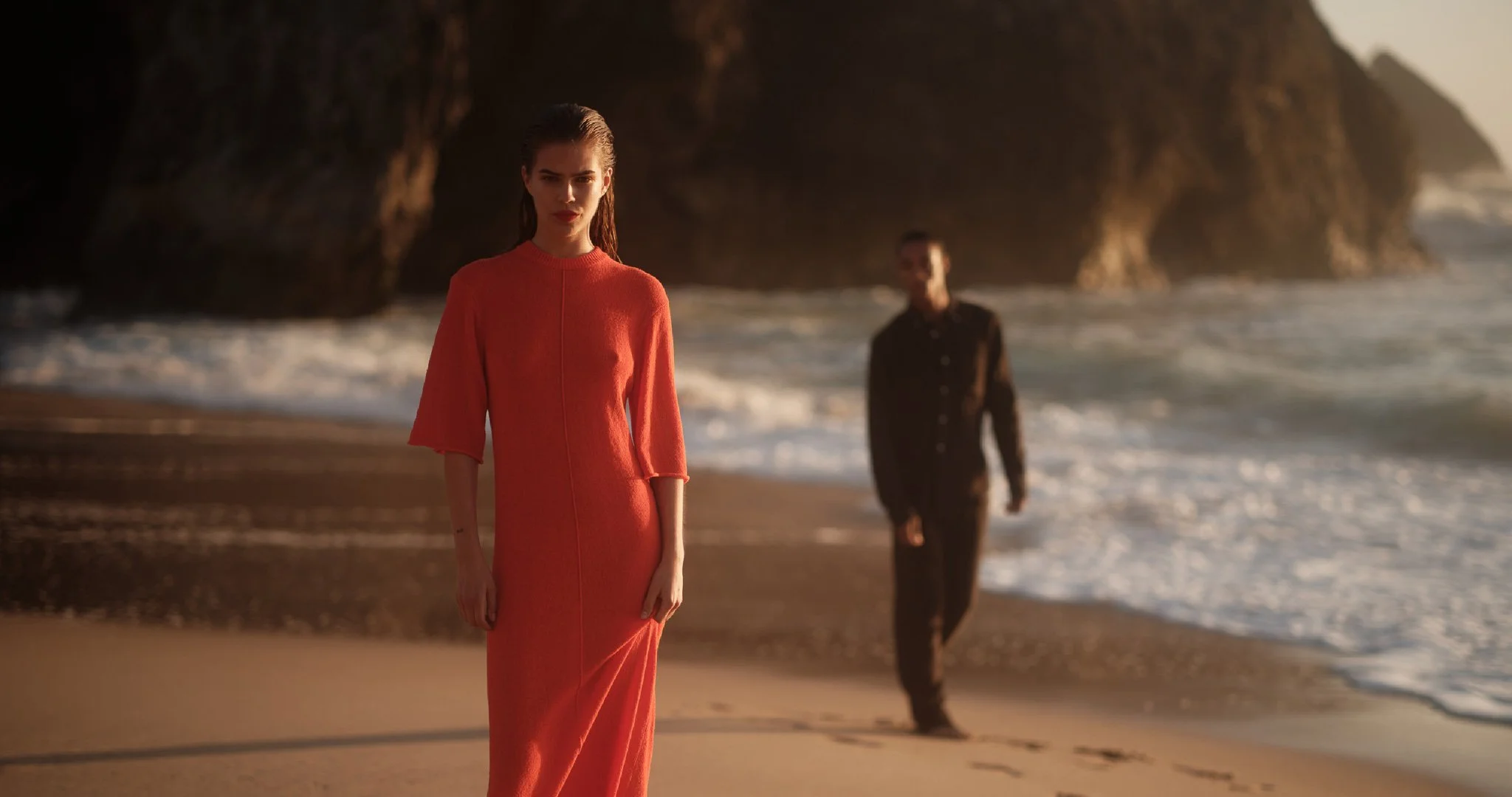 A woman in a coral-colored dress walking on a beach with a man in a black outfit behind her, near rocky cliffs during sunset.