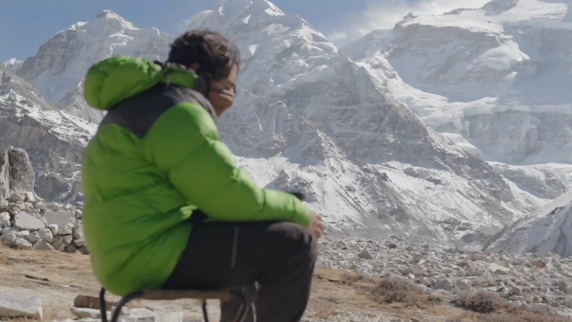 Person wearing a bright green jacket sitting on a small stool in front of snow-covered mountains