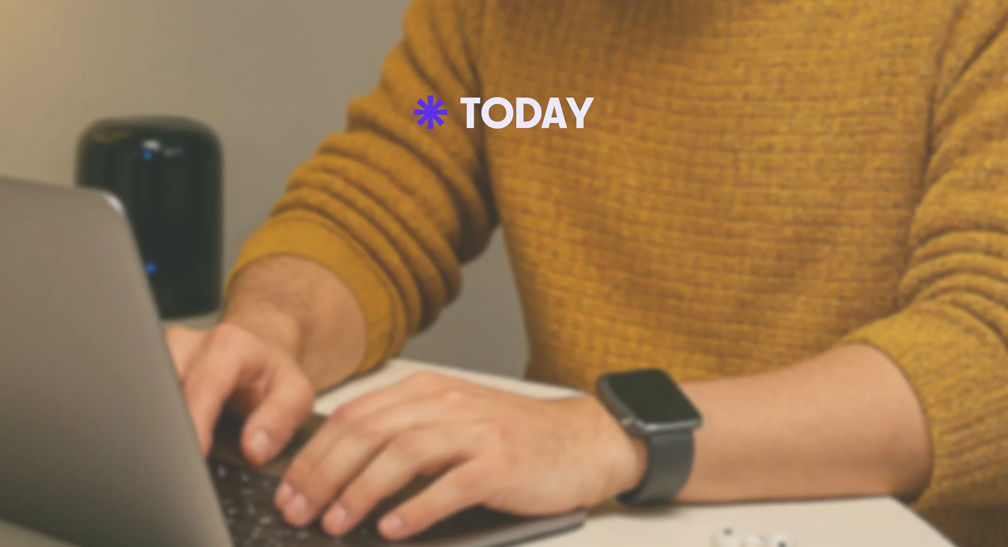 Person wearing a mustard yellow sweater and a black smartwatch typing on a laptop keyboard at a desk with a black speaker in the background.