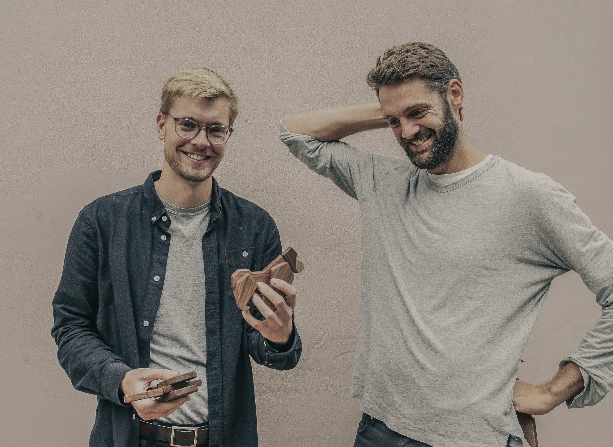 Two smiling men standing against a plain wall, one holding a wooden object and the other with his hand behind his head.