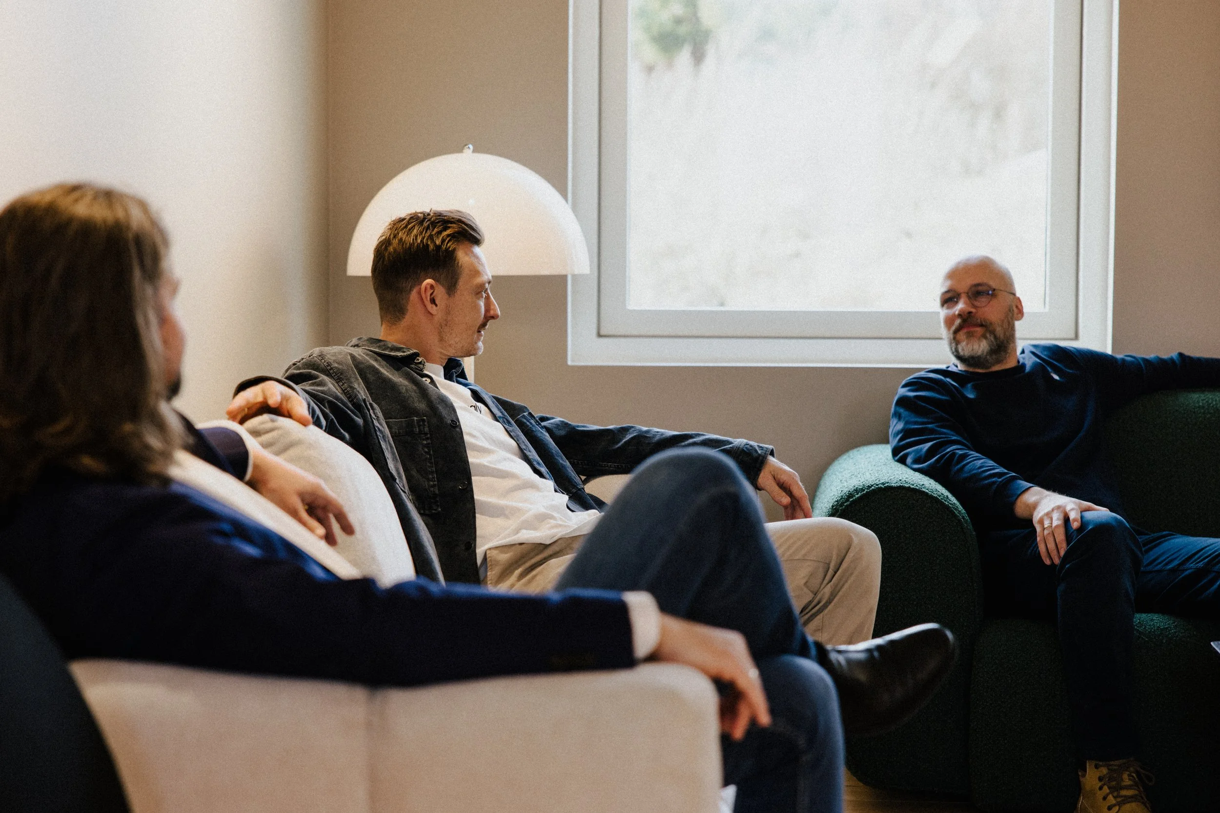 Four people sitting on couches in a casual indoor setting, having a conversation. The room has a large window and a white lamp.