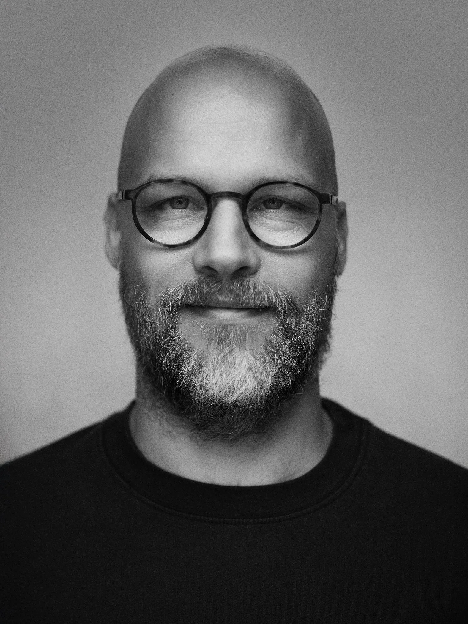 Black and white portrait of a man with glasses, a beard, and a bald head, smiling slightly and wearing a dark shirt.