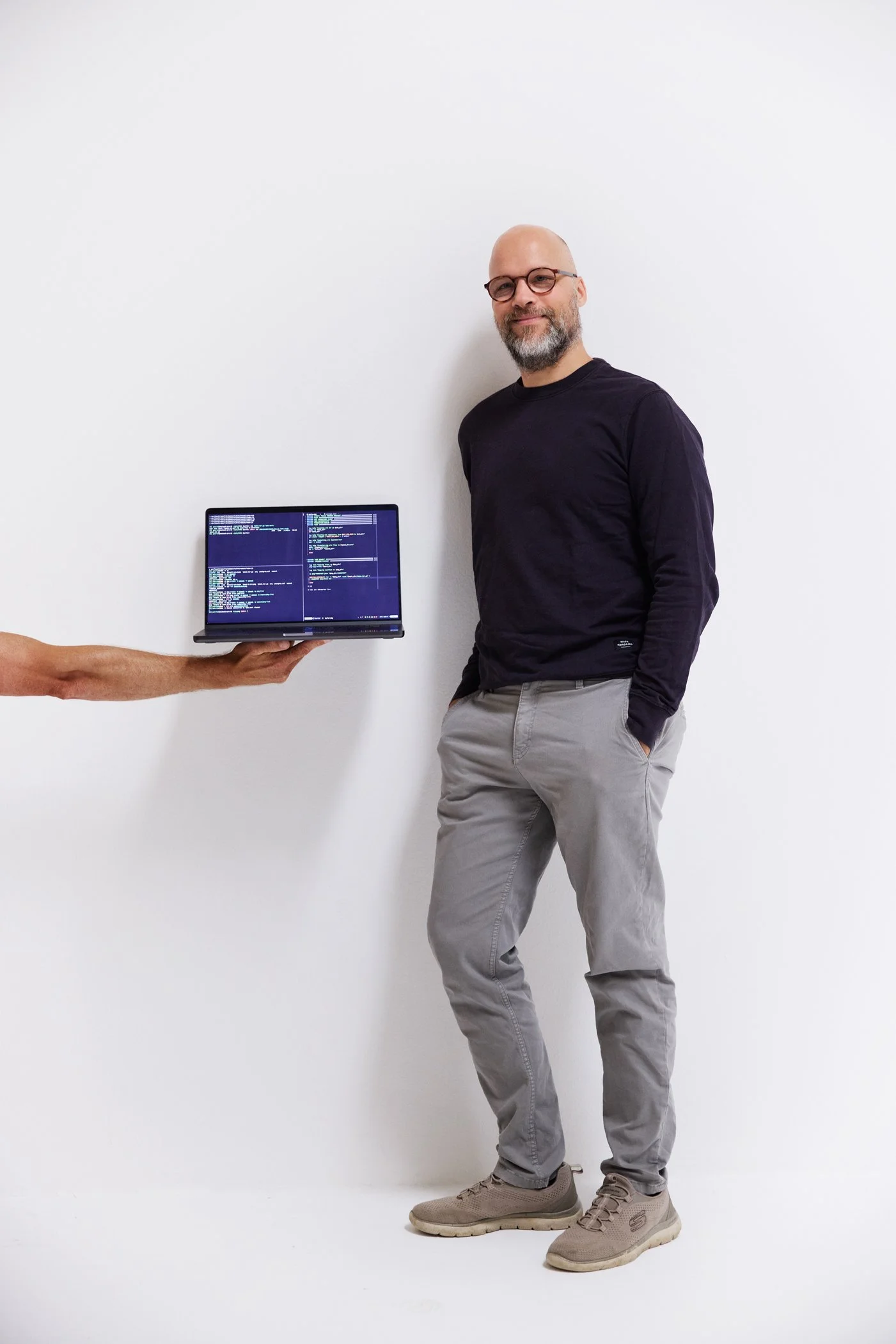 A man standing against a white wall, wearing glasses, a black long-sleeve shirt, gray pants, and beige shoes, while a person off-camera holds a laptop displaying coding or programming on the screen.
