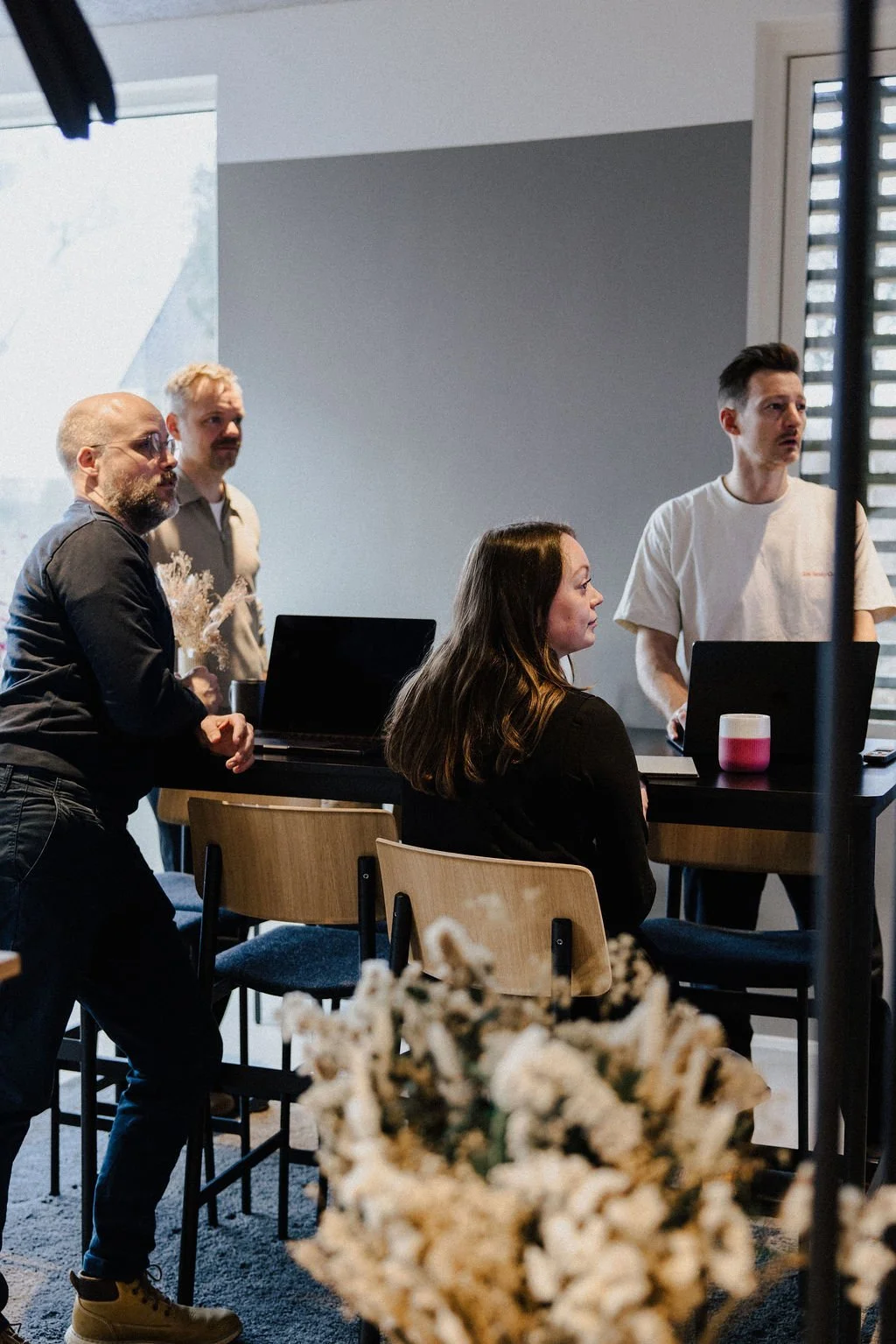 A group of five people, four men and one woman, in a modern room with laptops and a coffee cup, engaged in a discussion or presentation, with a blurred bouquet of flowers in the foreground.