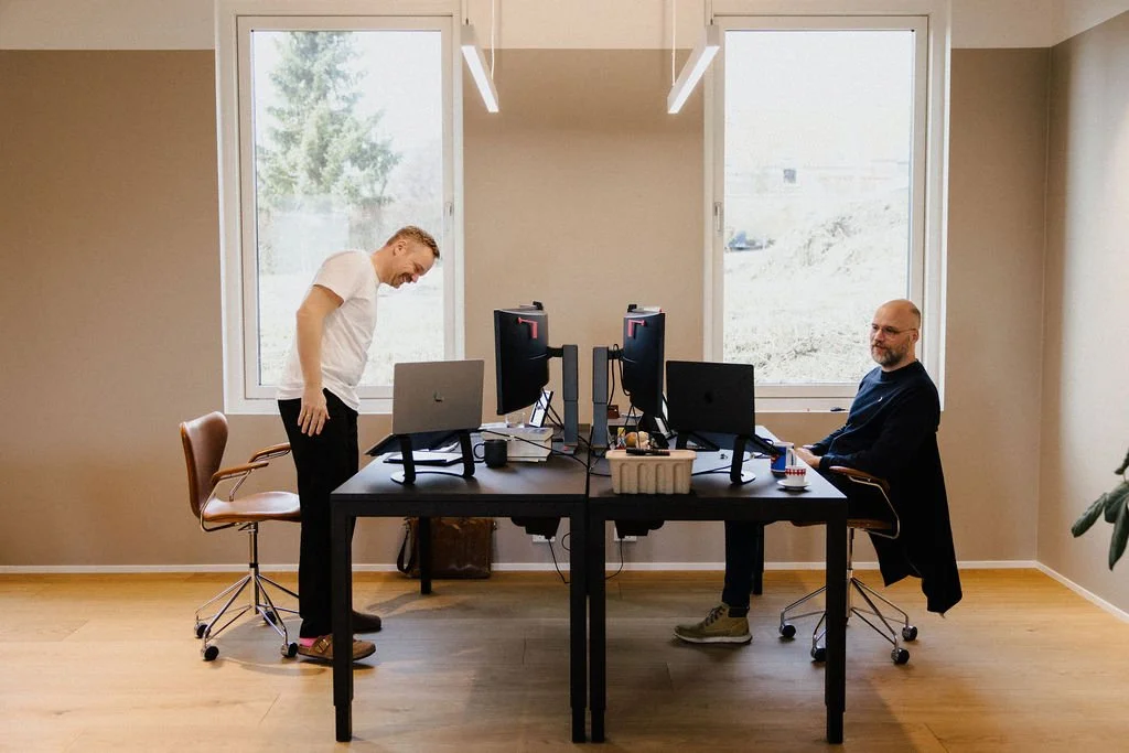Two men in a modern office with large windows, sitting at a black table with computer monitors, keyboard, and mouse. One man is smiling at the camera, while the other is looking down and leaning on the table. The office has wooden flooring and neutral-colored walls.