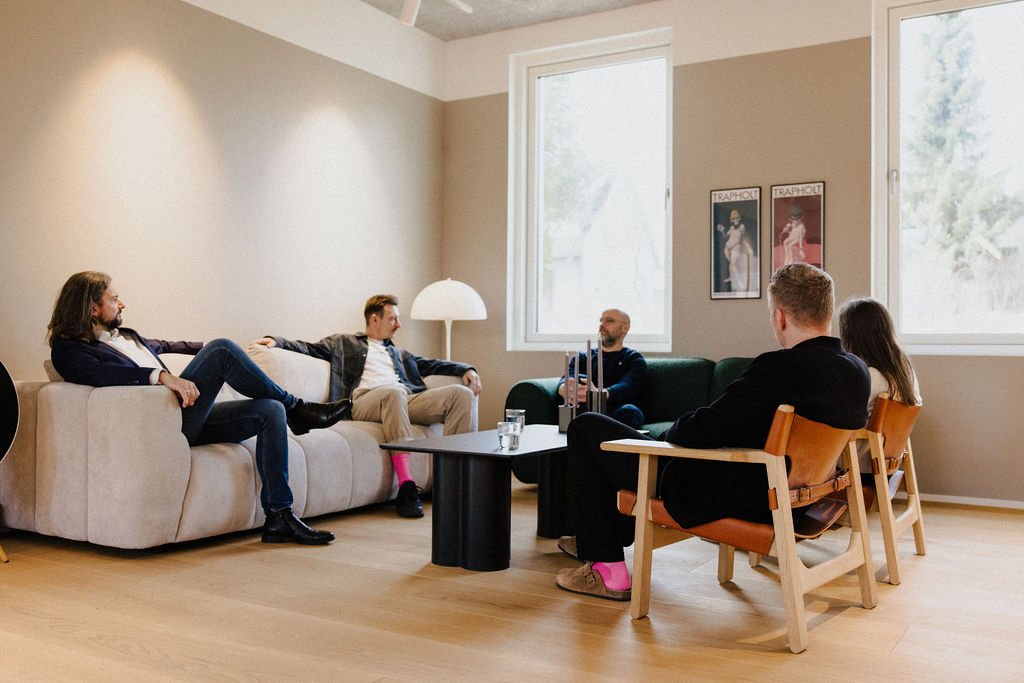 A group of six people sitting in a living room, engaged in a conversation. Four are seated on a beige sofa and two on wooden chairs with orange cushions. There are two large windows, a lamp, and framed artwork on the wall.