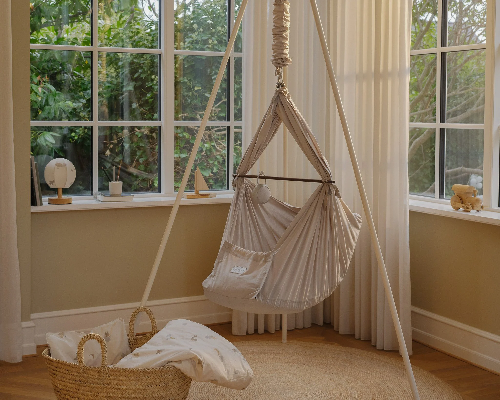 A cozy nursery corner featuring a beige baby hammock swing hanging from a wooden A-frame stand, with a wicker basket and a small pillow on a circular rug in front of it. The room has large windows with white curtains, decorated with small toys and ornaments on the window sills.