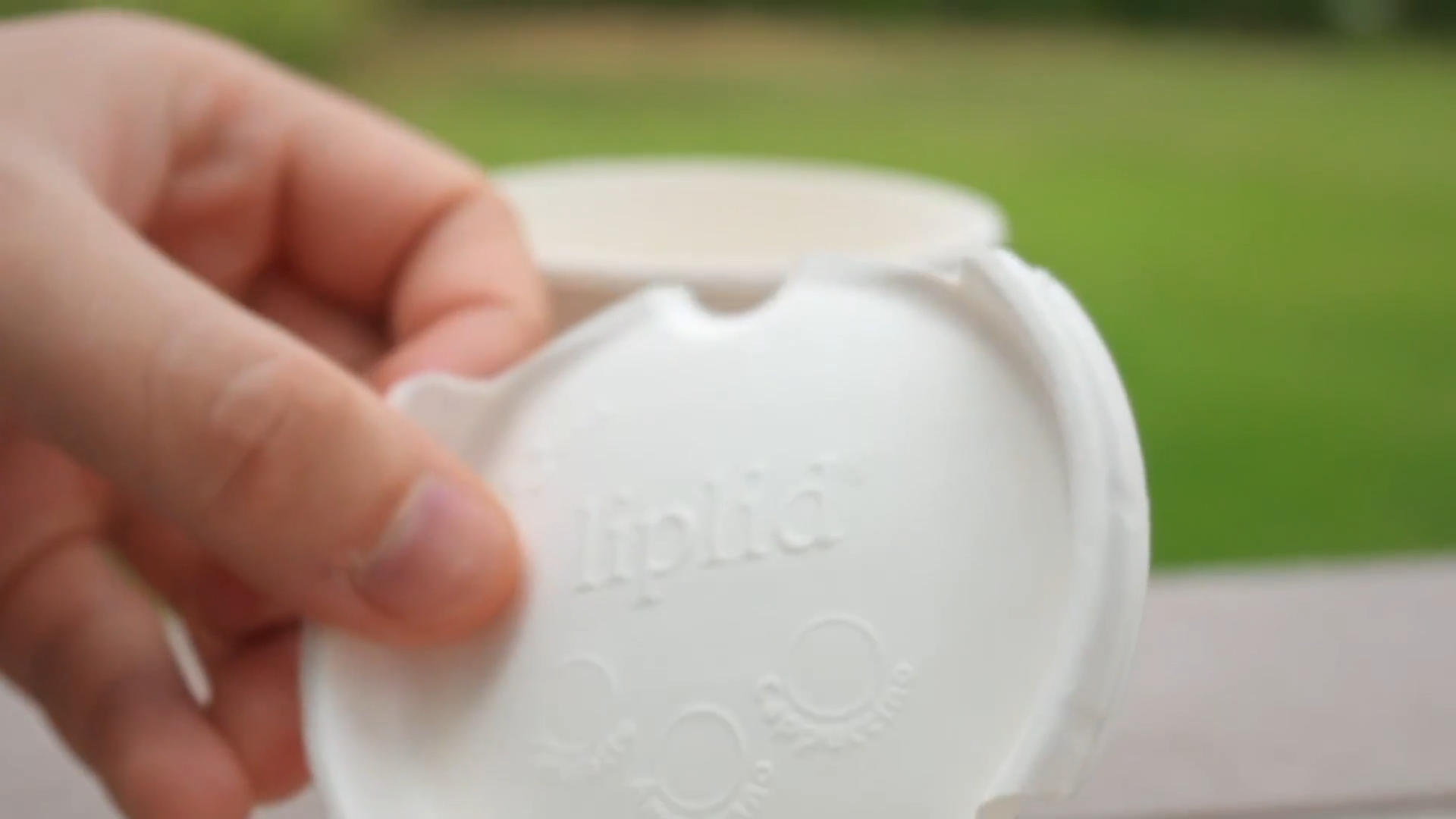 Close-up of a hand holding a white plastic container with an open lid, outdoors with blurred greenery in the background.