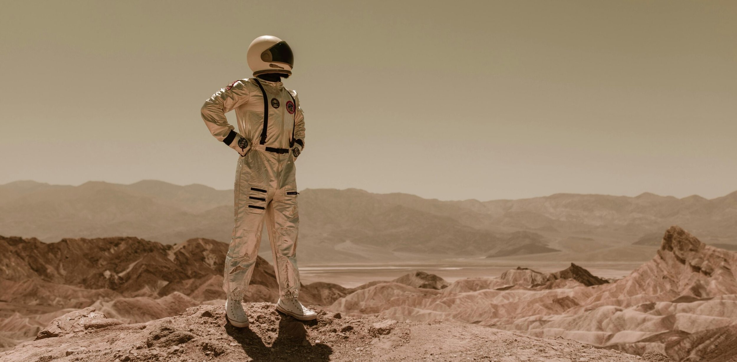 An astronaut in a silver spacesuit standing on rocky terrain with mountains in the distance, under a hazy sky.