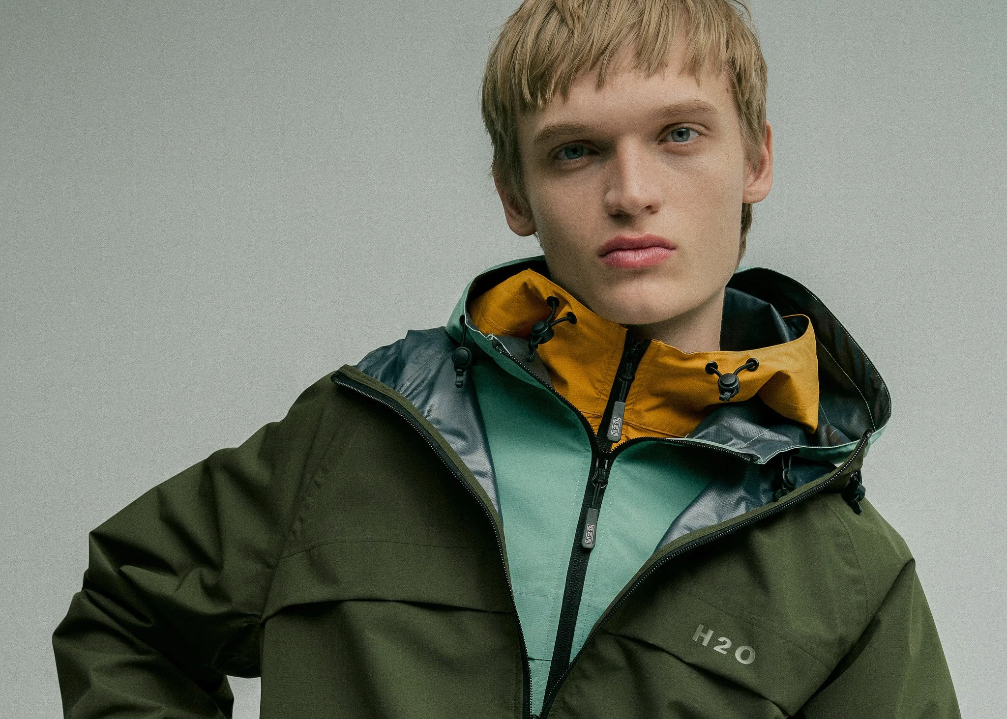 A young man with blond hair and blue eyes wearing a green and black waterproof outdoor jacket, standing against a plain gray background.