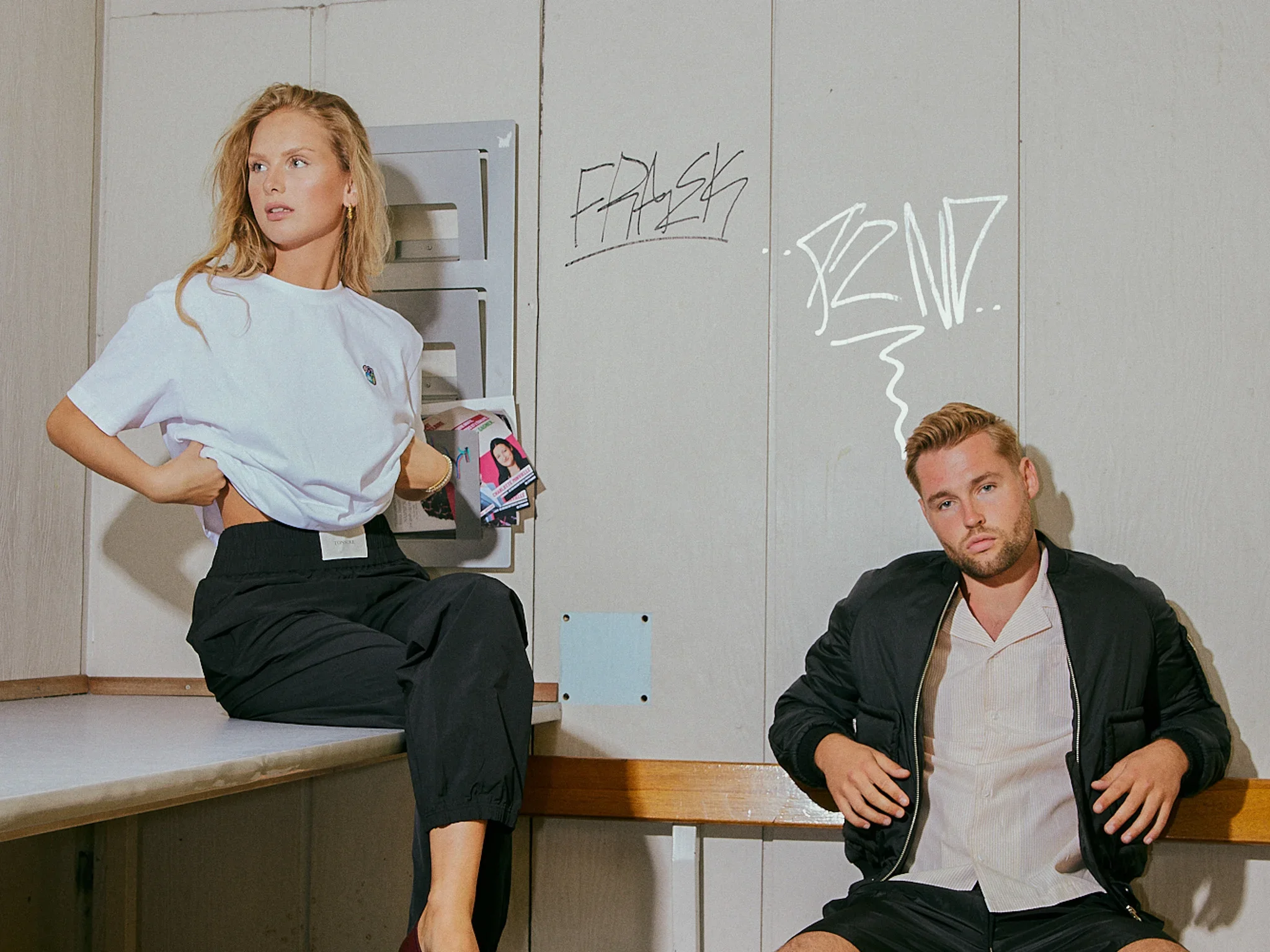 A woman sitting on a counter with hands behind her back, and a man sitting on the floor with head tilted, wearing a black jacket and white shirt, in a room with graffiti on the wall.