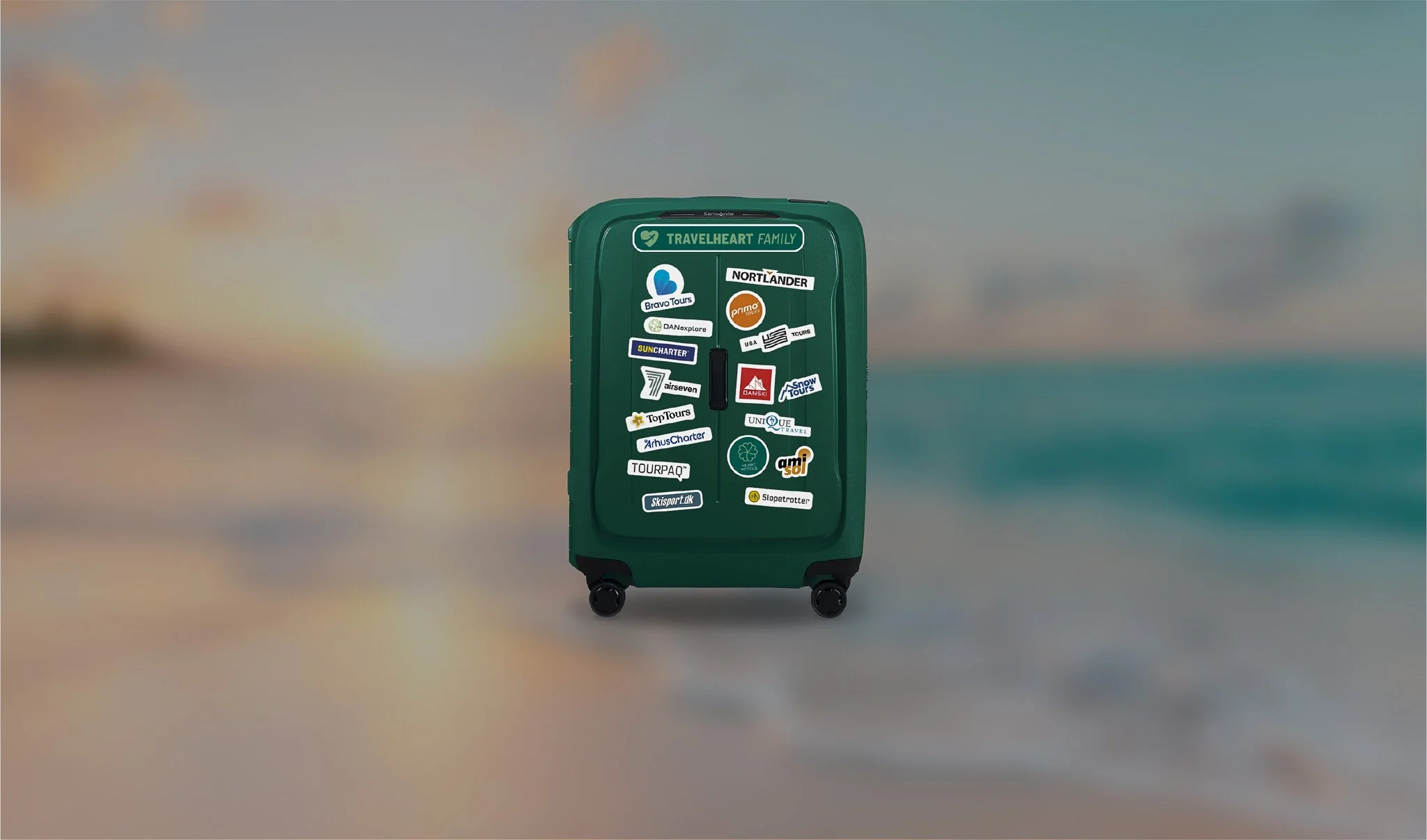 Green suitcase covered with travel-related stickers standing on a beach with blurred sand, ocean, and sky in the background.