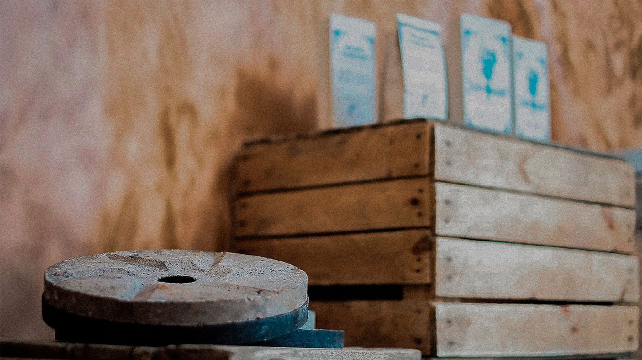 Close-up of a rustic, round, metal object with a hole in the center, in front of stacked wooden crates and blue instructional posters on a textured wall.
