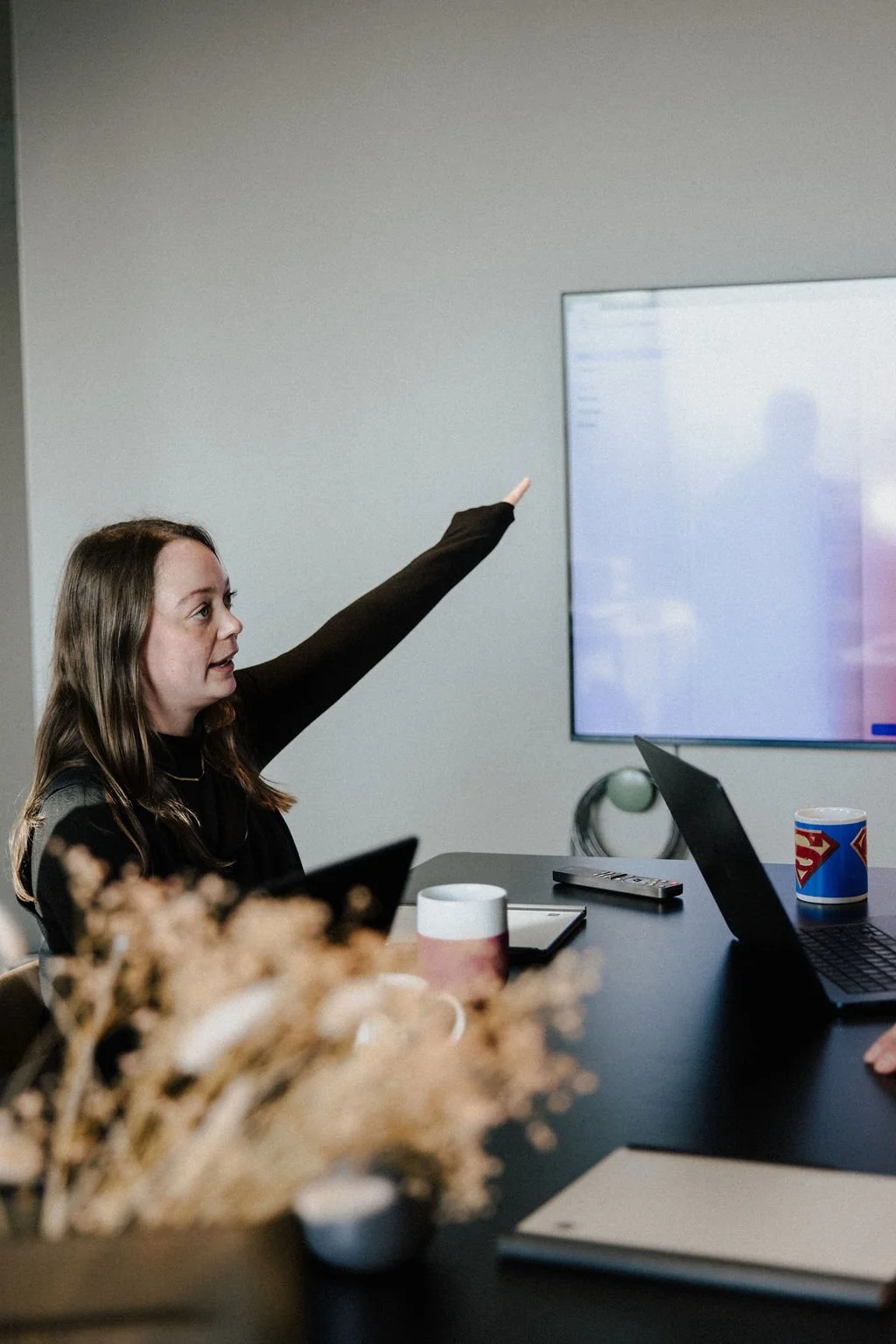 A woman is sitting at a conference table, raising her hand to point at a presentation on a screen in a meeting room.