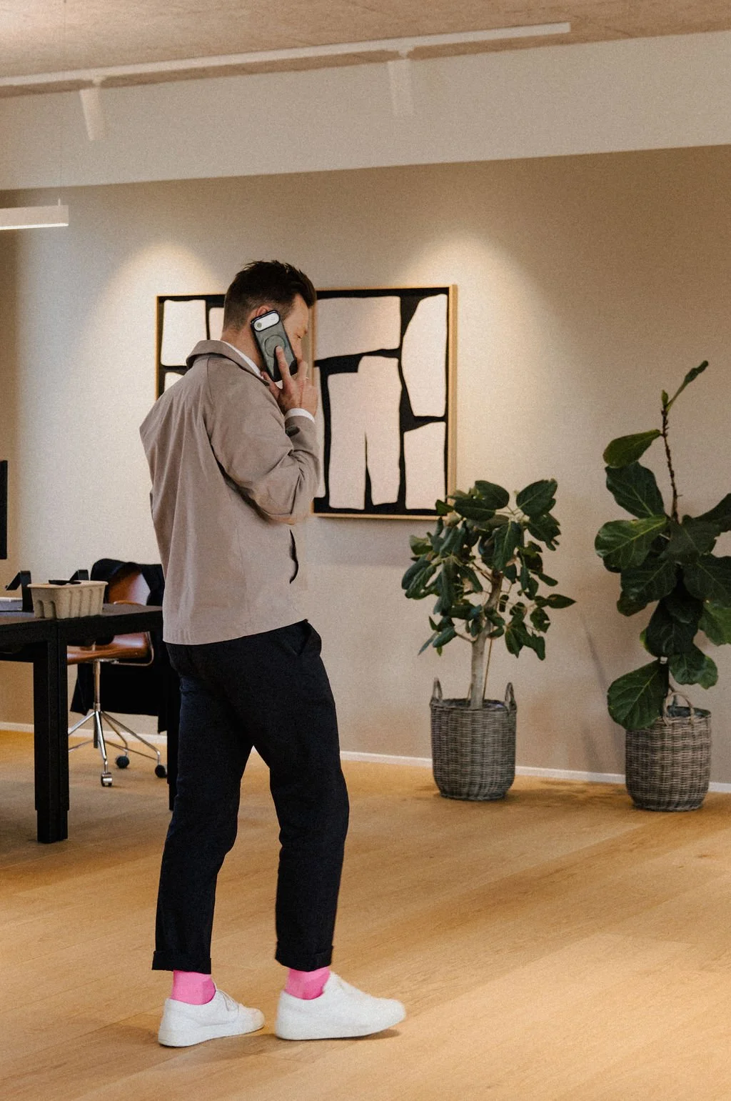 A man in a beige jacket, black pants, white sneakers, and pink socks is talking on a cell phone in an office with potted plants and modern abstract artwork on the wall.