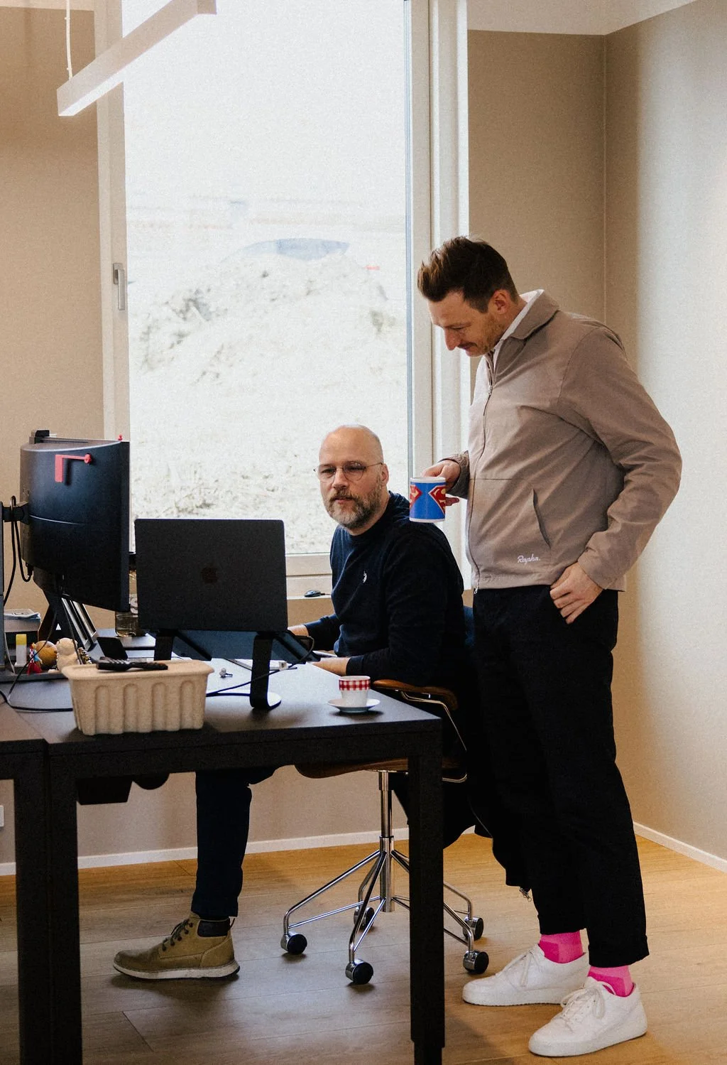 Two men in a home office, one sitting at a desk working on a computer and the other standing beside him, holding a colorful cup, with a large window and a view of a gravel area outside.