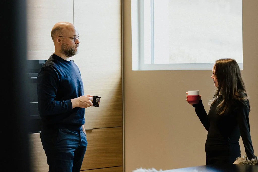 A man and a woman are standing indoors, holding coffee mugs, and facing each other in a well-lit room with a large window.