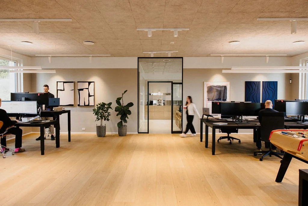 Modern office space with desks, computers, and employees working. There is a glass partition leading to a conference room, and abstract art on the walls.