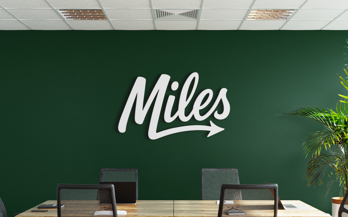 Conference room with a green wall, a large 'Miles' sign written in white with a right-pointing arrow, and a wooden table with black mesh chairs, with a potted plant on the right side.