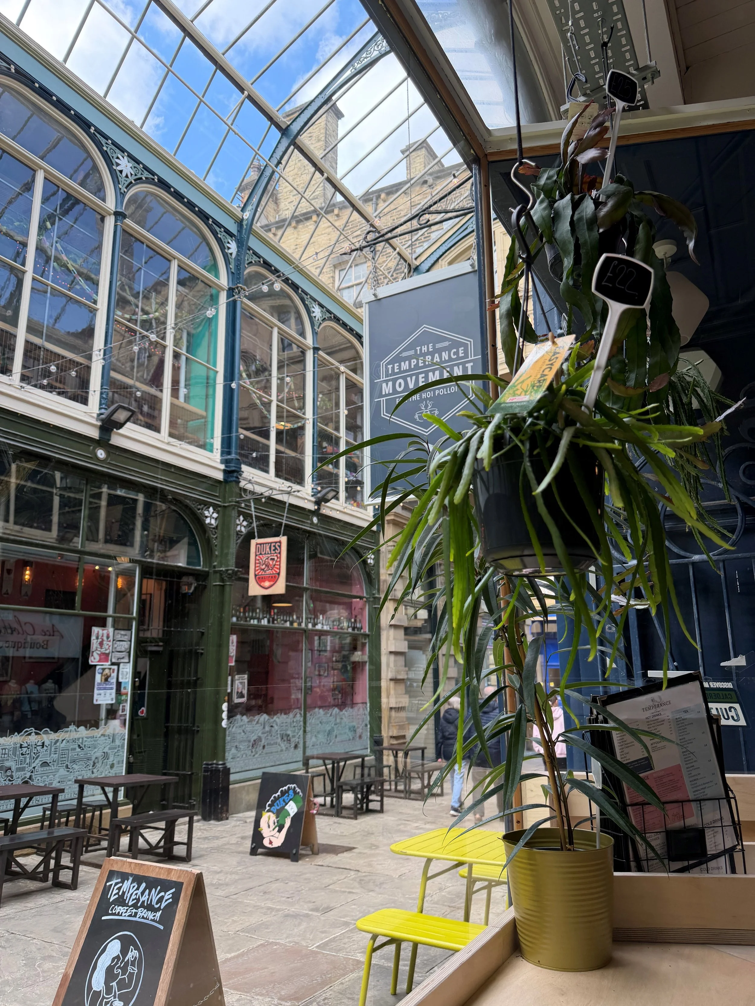 Temperance Coffee Bar, Market Street in Halifax. Indoor market or cafe with hanging plants, chalkboard signs, and a view of an outdoor courtyard with tables and storefronts.