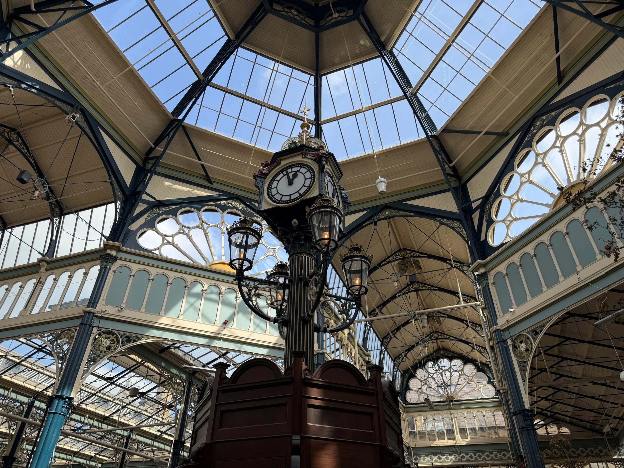 Interior of a historic Halifax Market with a glass ceiling, ornate ironwork, and a central clock showing the time as 2:50.