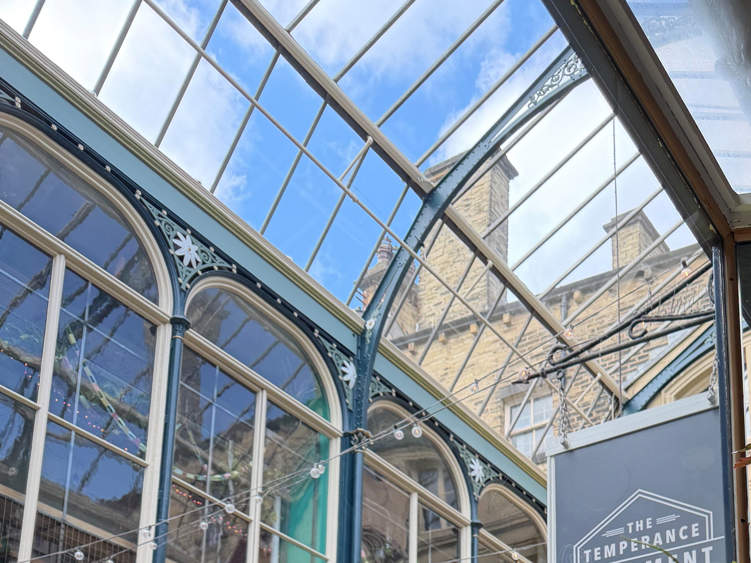 Interior view of a glass ceiling with a blue sky and clouds visible, parts of an ornate building with stone chimney stacks, and a sign reading 'The Temperance' inside a historic or vintage structure.