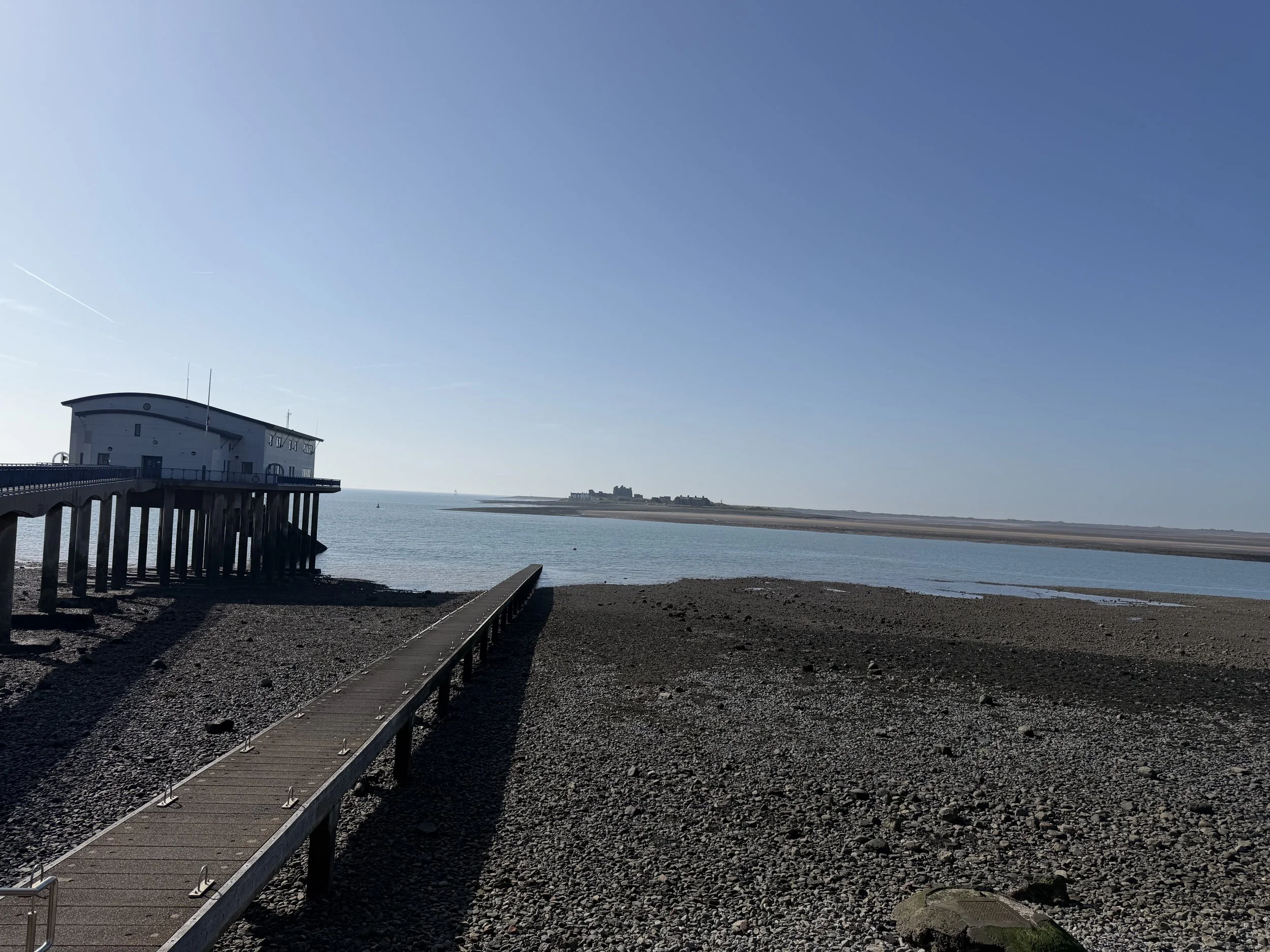 View across to Piel Island, Cumbria. A pier extending into a calm body of water near a rocky shore with a building on stilts at the pier's end and land with a castle-like structure in the distance under a clear blue sky.