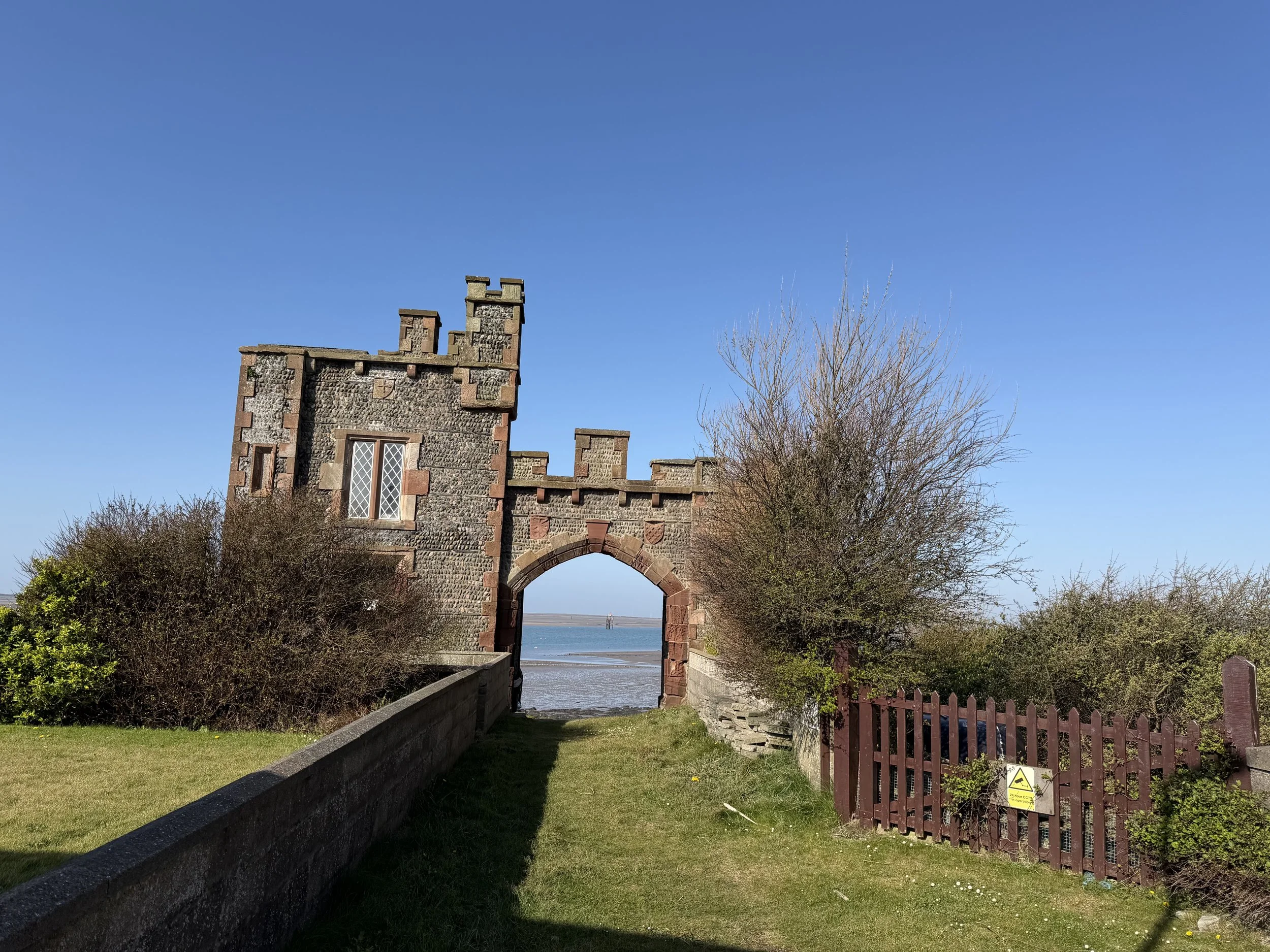 Piel Island, near Barrow-in-Furness. Old stone castle-like structure with an archway, overlooking a body of water, with bushes and trees on either side, under a clear blue sky.
