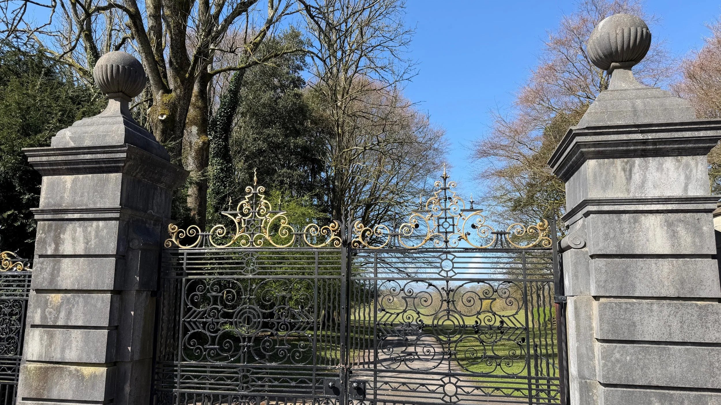 Holker Hall stone gate with ornate black and gold ironwork, flanked by large stone pillars topped with decorative stone urns, leading to a park or garden area with trees and a pathway.
