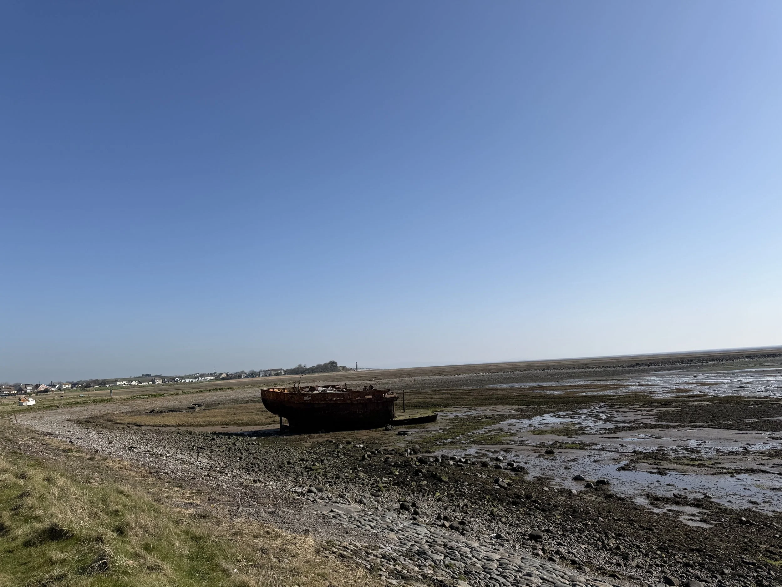 Old wooden boat stranded on the muddy shoreline of a tidal flat under a clear blue sky, with a small village visible in the background. Bardsea Cumbria
